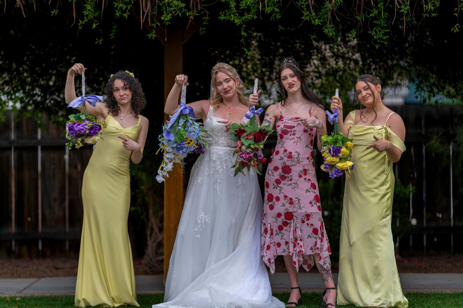 Bride in white gown poses with three bridesmaids in yellow dresses holding colorful bouquets outdoors.