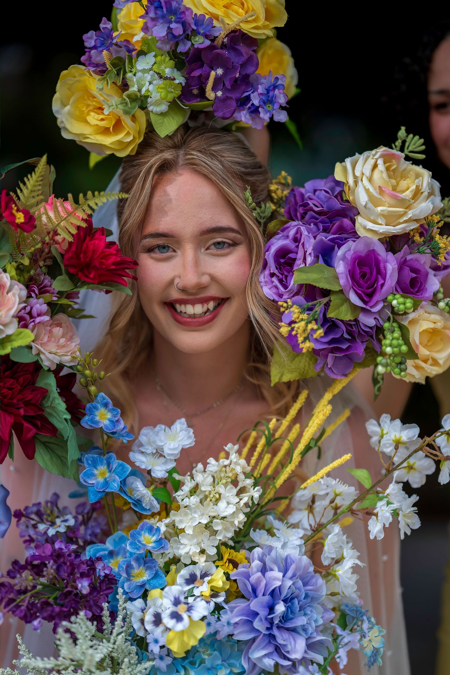 A smiling bride surrounded by vibrant colorful wedding bouquets featuring purple, yellow, and blue flowers.
