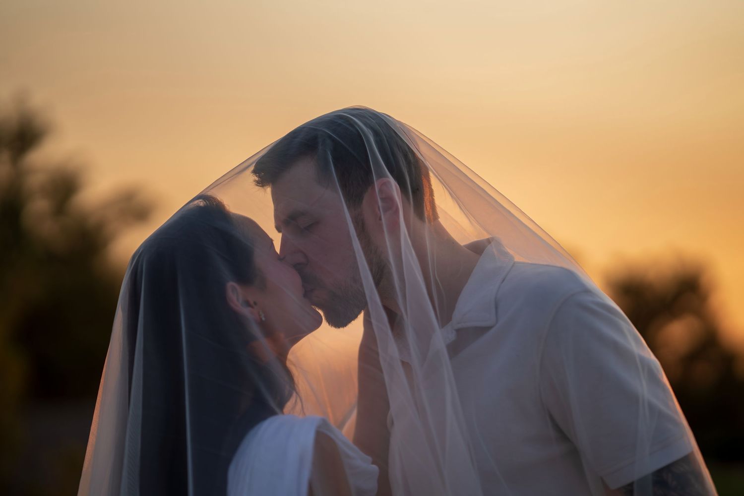 A bride and groom share a romantic kiss under a wedding veil at golden hour sunset.