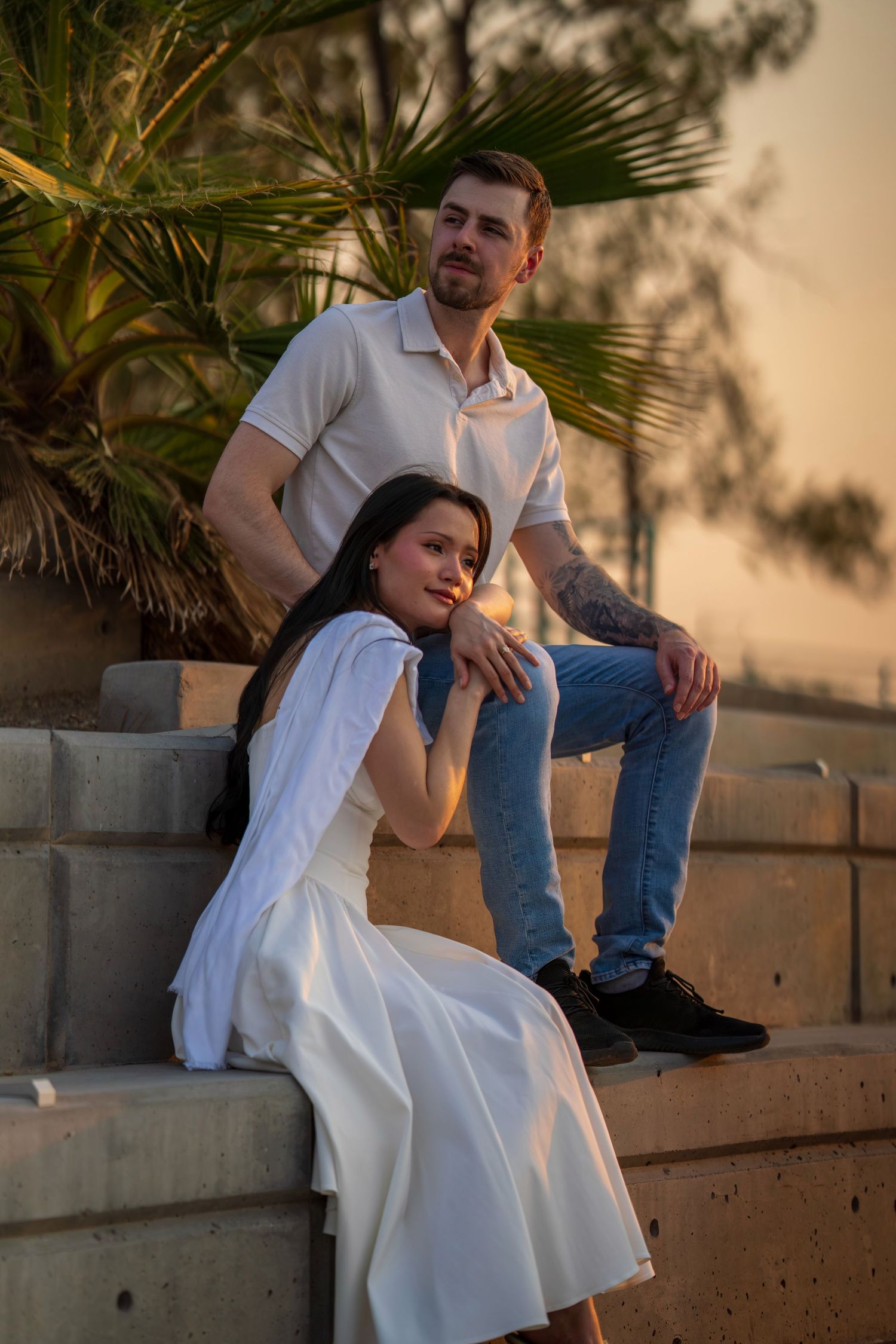 Stylish couple posing on concrete steps at sunset, woman in white dress and man in casual white shirt and jeans.
