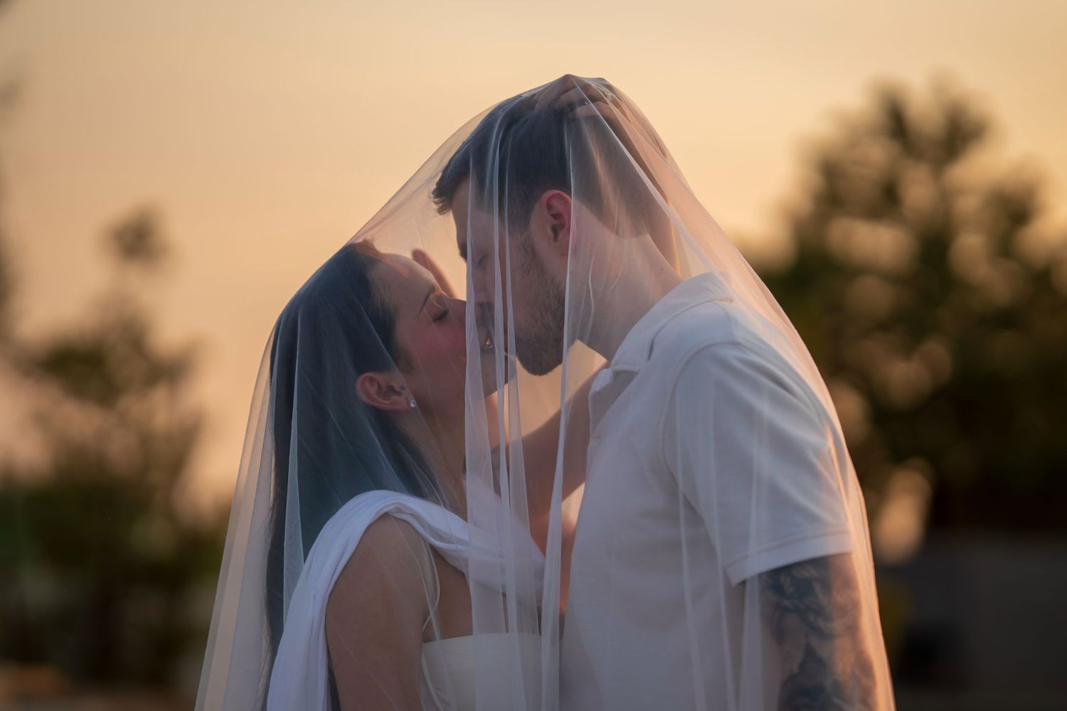 Newlywed couple sharing kiss under bridal veil during golden sunset with trees in background.