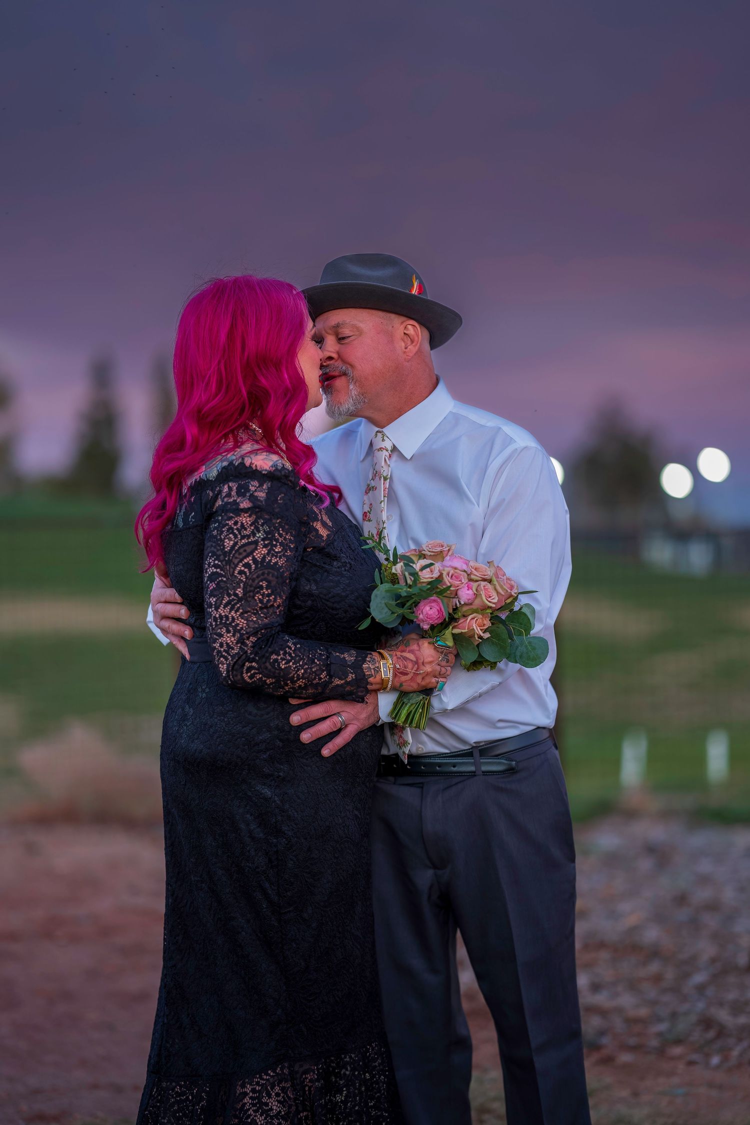 Couple embracing at sunset, woman with pink hair in black lace dress holding pink bouquet, man in white shirt and gray hat.