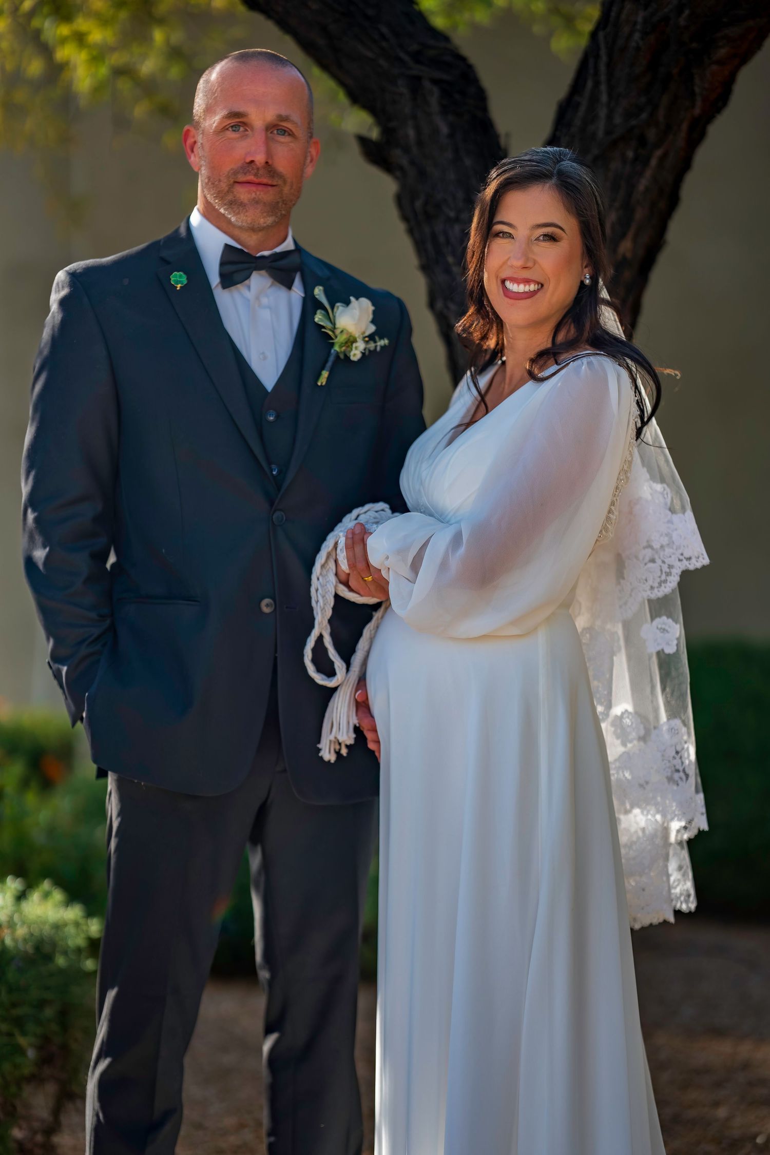 Bride and groom pose formally for portrait under tree, bride holding lace-trimmed bouquet at outdoor wedding.