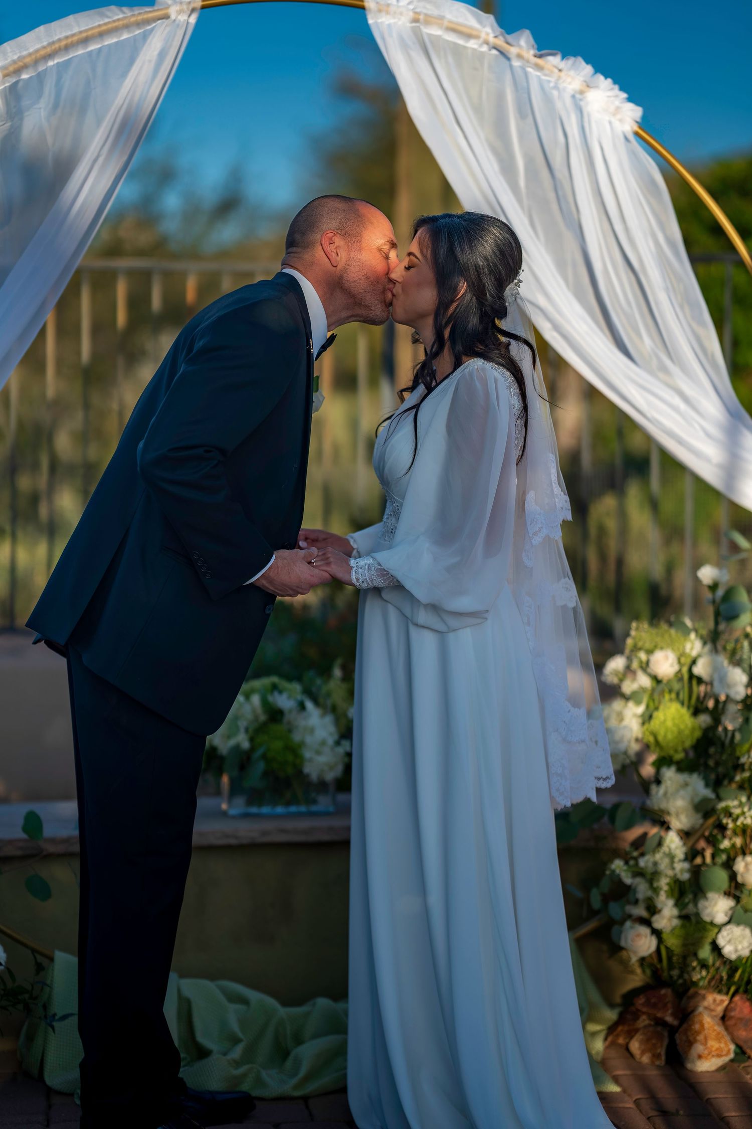 Bride and groom sharing first kiss under a white draped arch adorned with white floral arrangements.