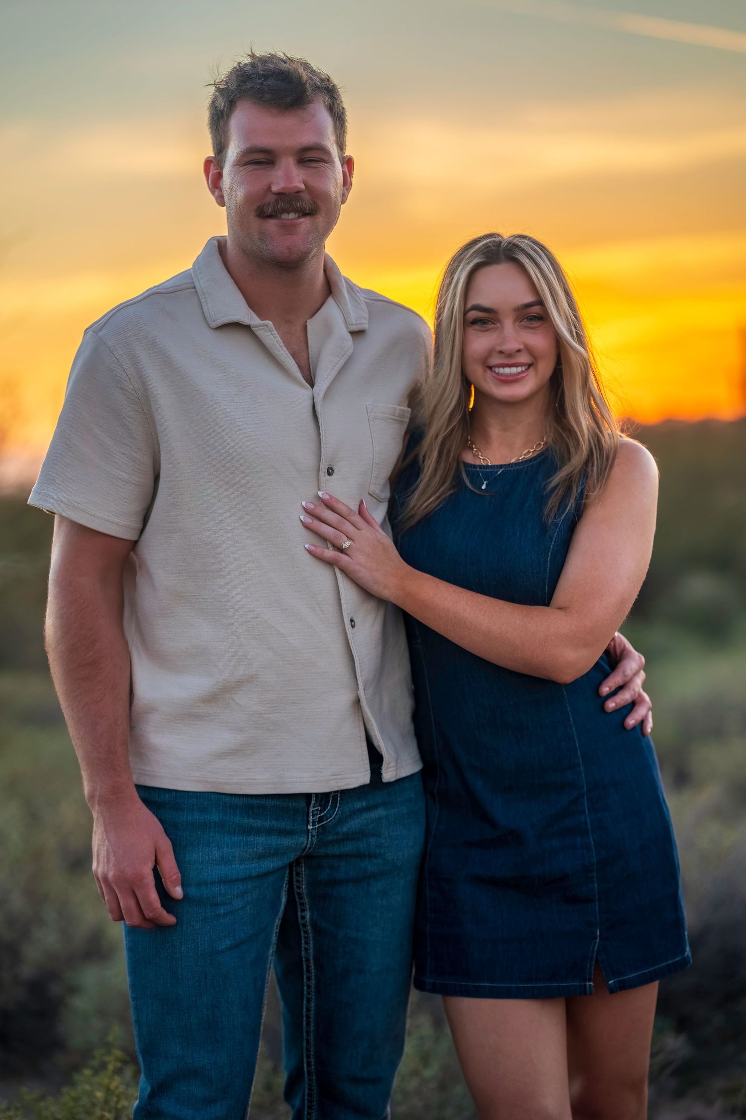 Couple posing at sunset in desert landscape, man in beige shirt and jeans, woman in navy blue dress smiling.