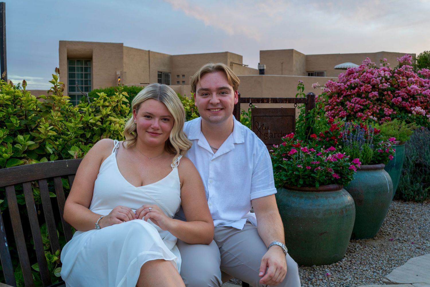 Newly engaged couple sitting together on a bench surrounded by lush garden flowers at dusk.