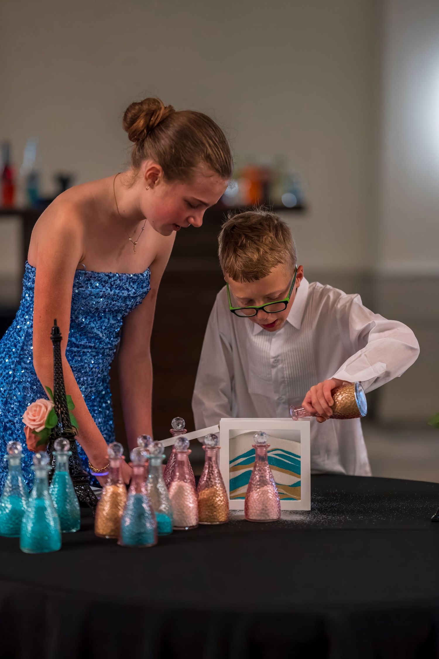 Girl in blue sequin dress and boy in white shirt pour colored sand into a frame at a wedding ceremony.