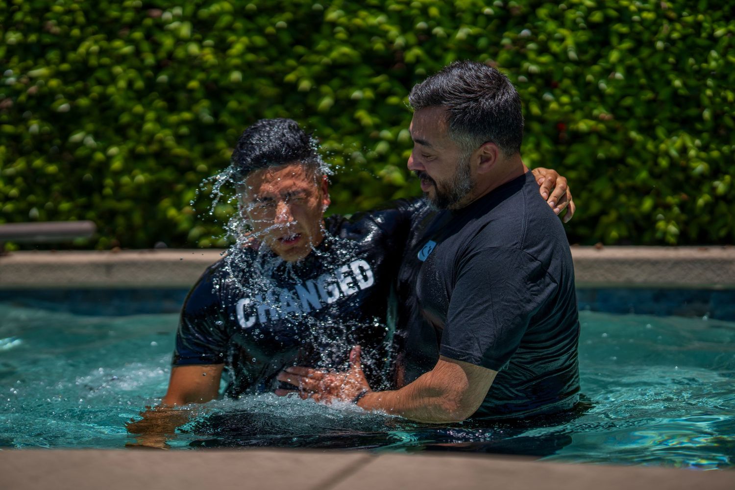 Two men in a pool during a baptism ceremony, one wearing a 'Changed' shirt being immersed in water.
