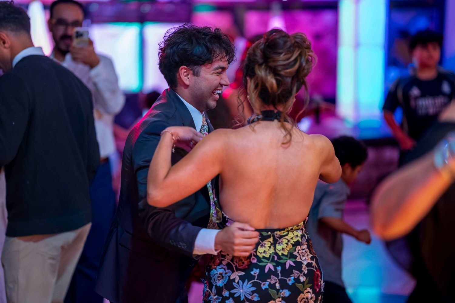 Couple dancing closely at a vibrant party, man in suit smiling at woman in floral backless dress.