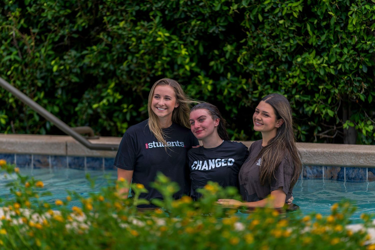 Three smiling young women standing together in an outdoor pool wearing matching church ministry t-shirts.