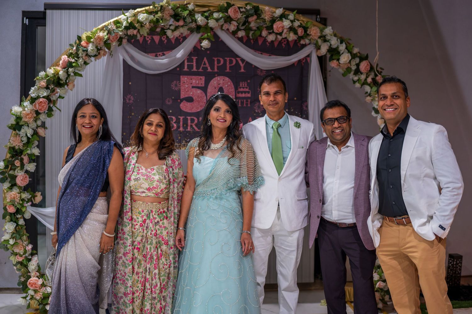 Guests in formal wear gather for photos under an ornate floral arch at a celebration.