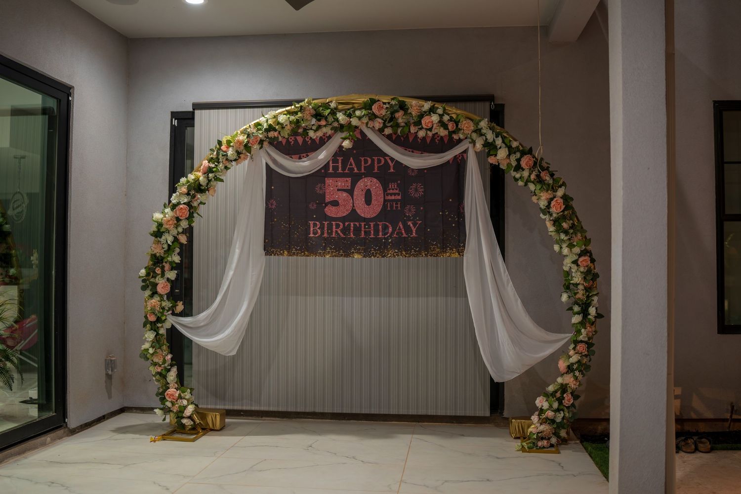 Elegant party venue with floral arch, white drapes and birthday celebration backdrop.