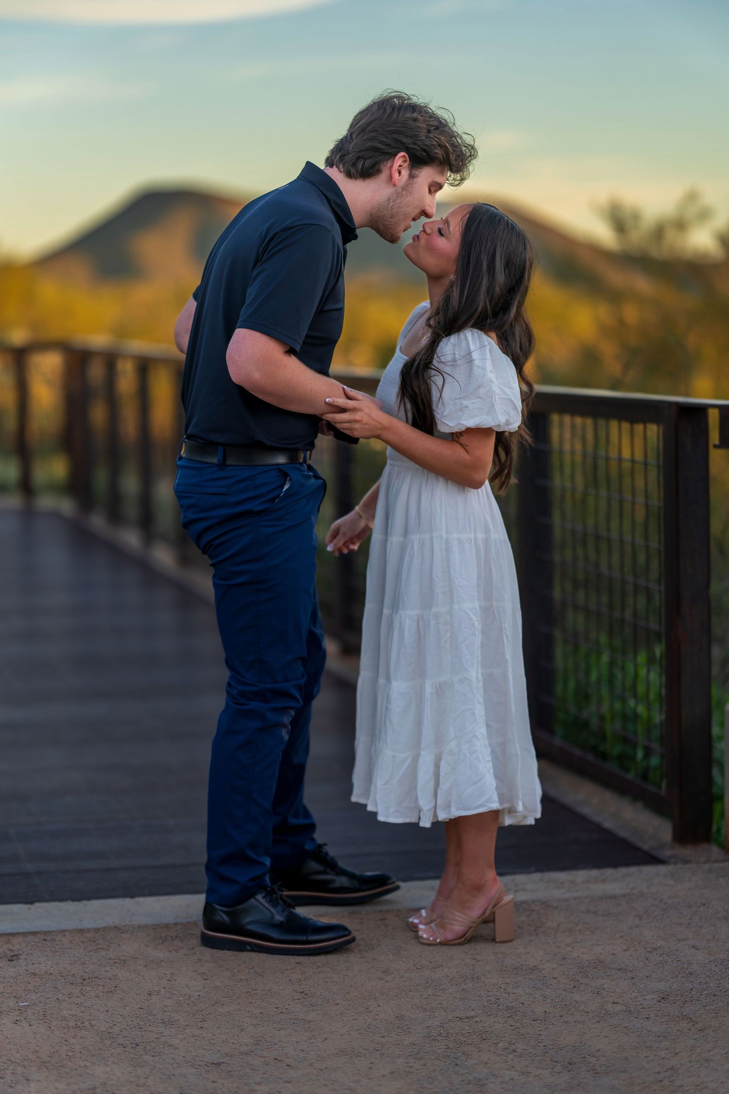 Two people share a tender moment on a desert viewing platform with mountains in the background.