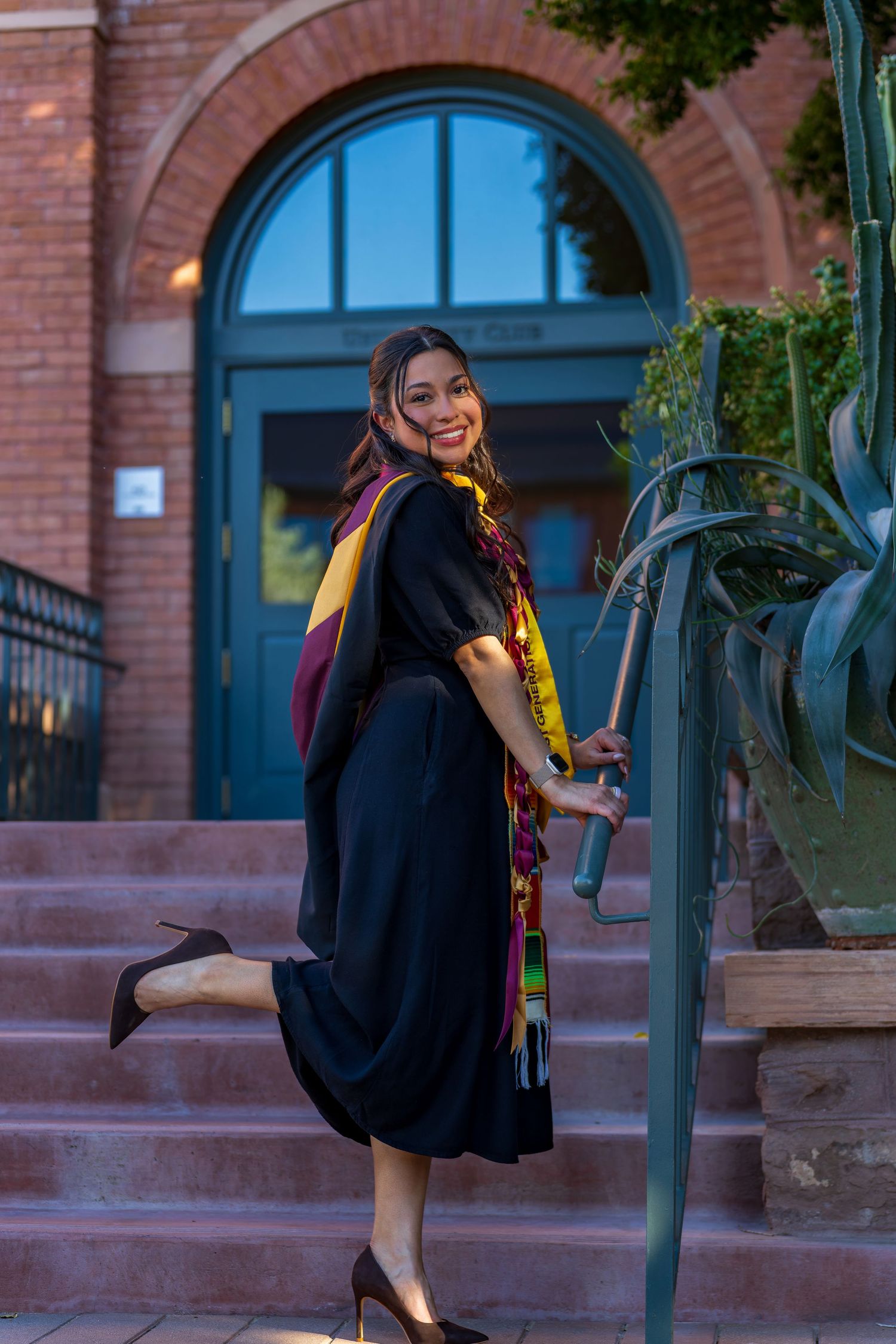 Graduate in black dress and colorful stole poses on brick building steps at dusk.