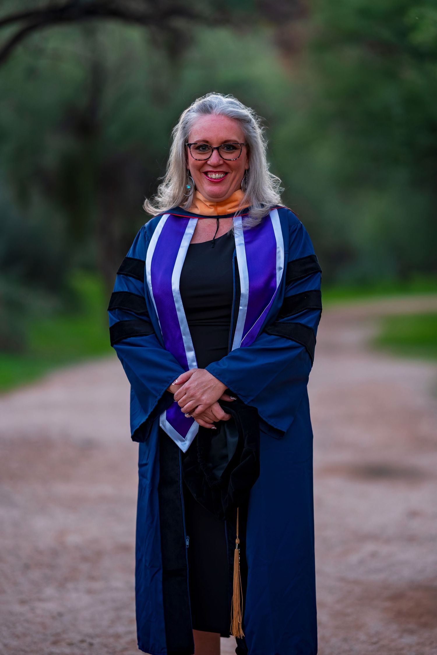 Graduate in blue academic regalia with purple stole stands smiling on outdoor path.