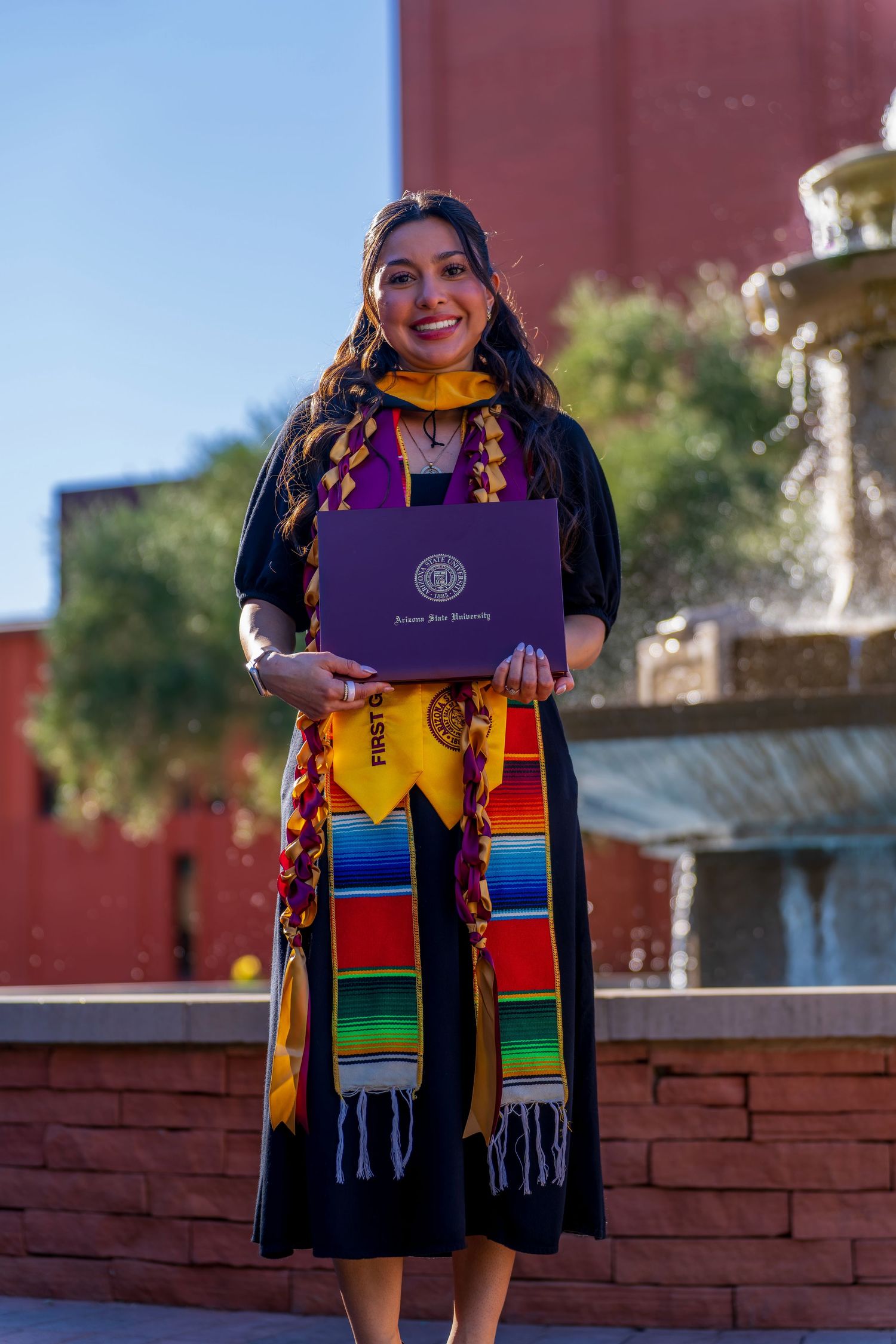Graduate in ceremonial dress holds diploma while standing by brick wall.