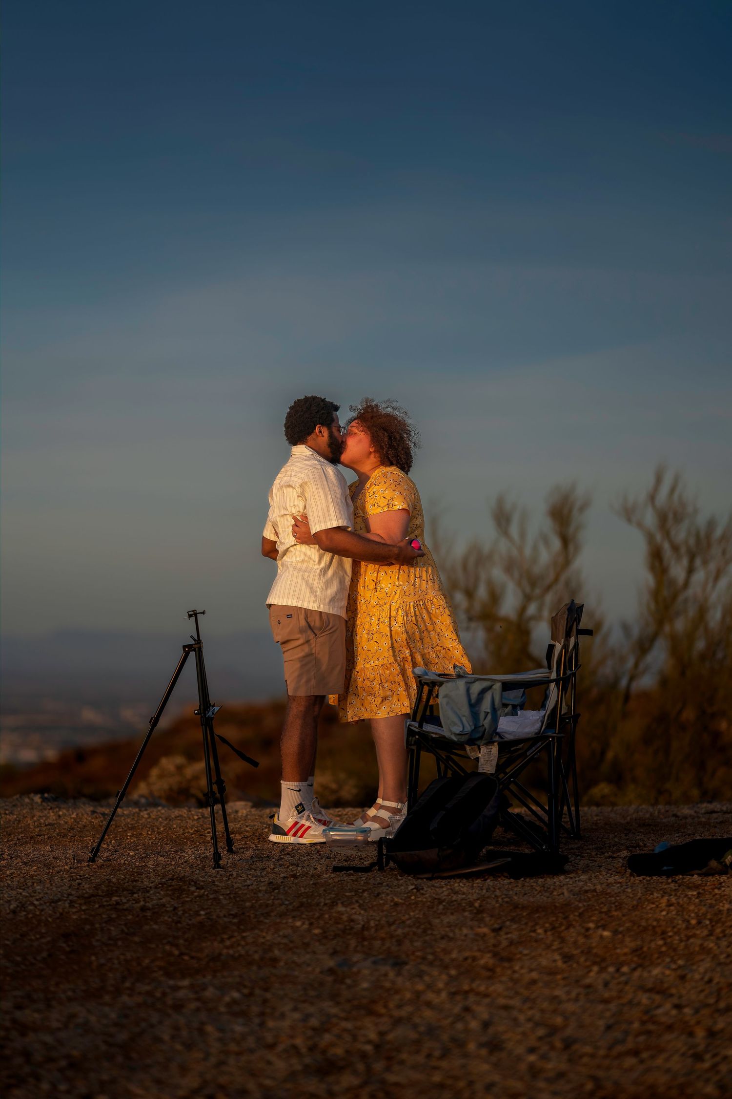 Two people embracing and kissing during golden hour on a scenic overlook with desert shrubs and a tripod nearby.