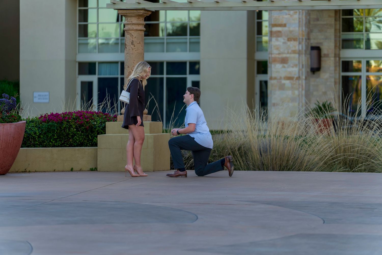 Man proposing on one knee to woman outdoors near modern building entrance with stone pillars and pink flowers.
