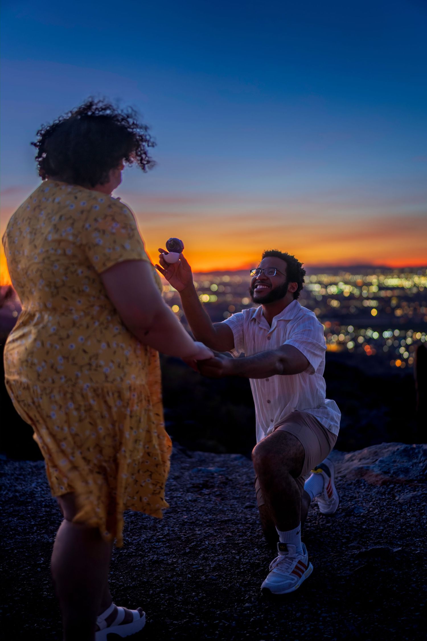 Joyful marriage proposal at twilight with man on one knee holding ring box, city lights behind.