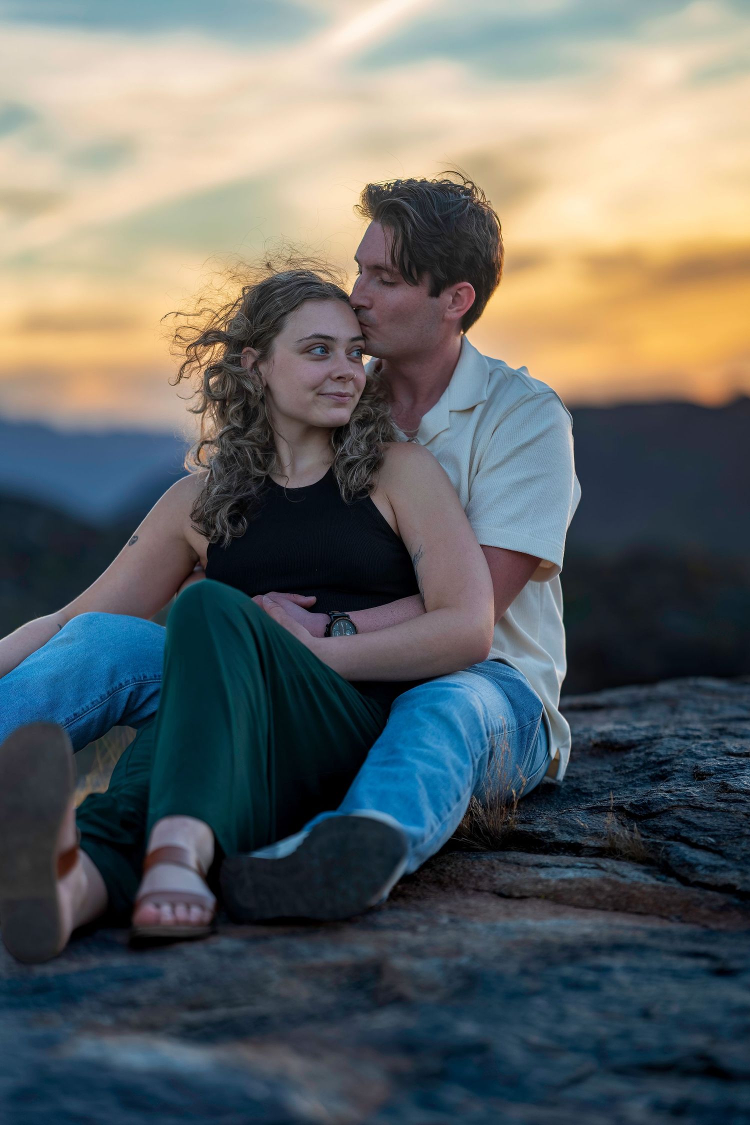 Couple sitting on a rocky overlook embracing at sunset with mountain views.