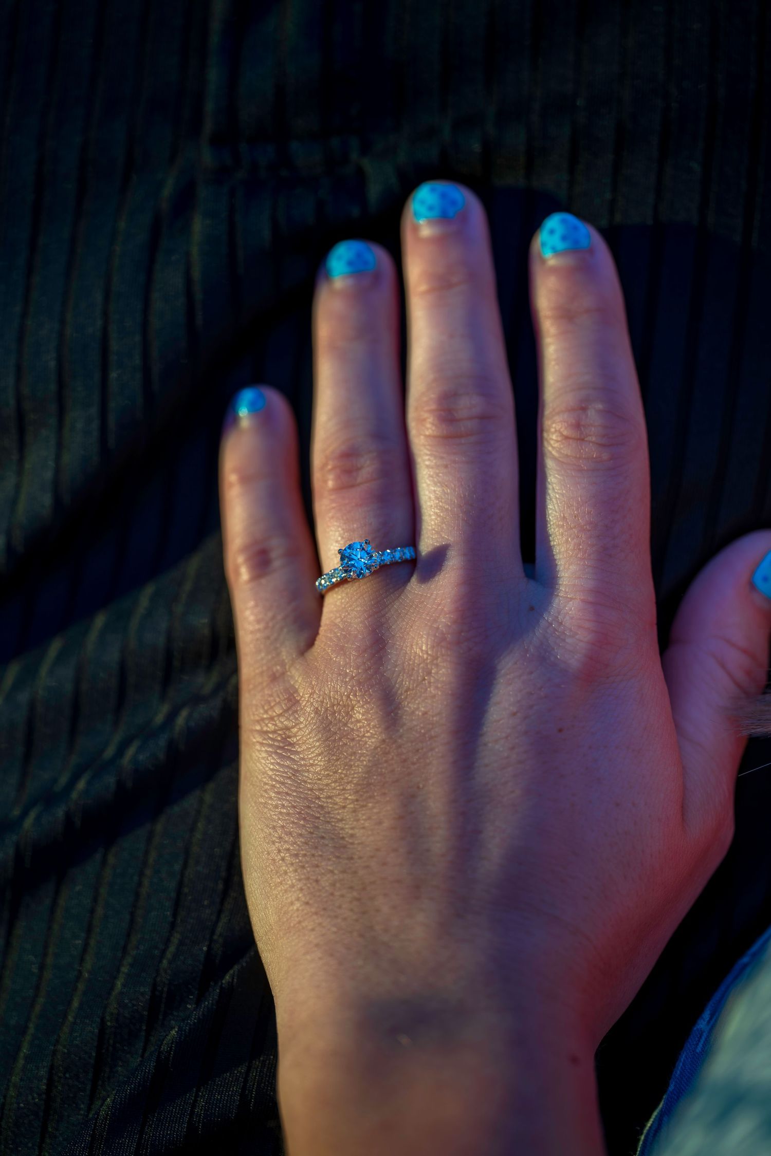 Close-up of engagement ring with blue gemstone on womans hand with blue painted nails resting on dark fabric.