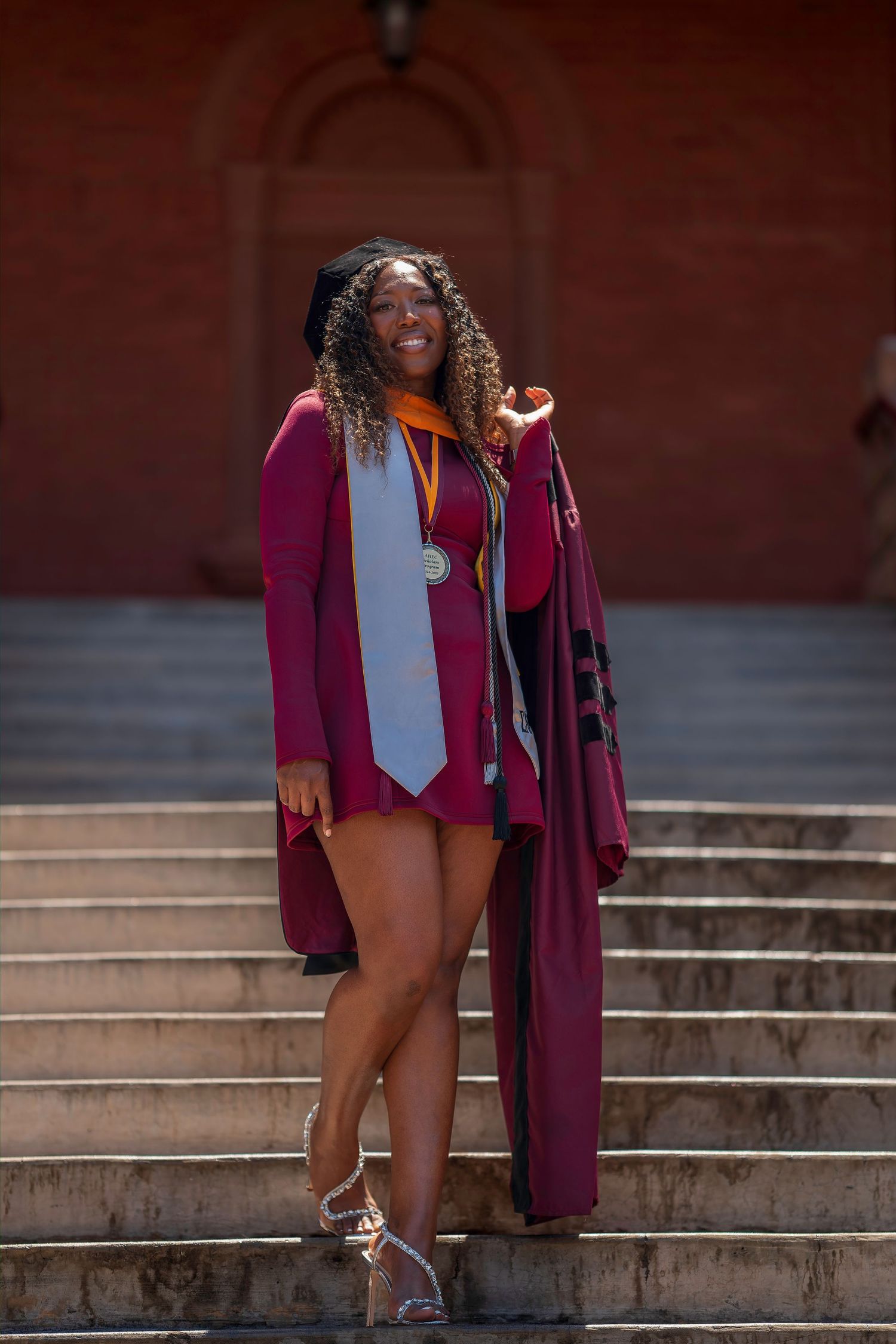 Young woman in maroon graduation gown and stole poses proudly on stone steps, smiling on a sunny day.