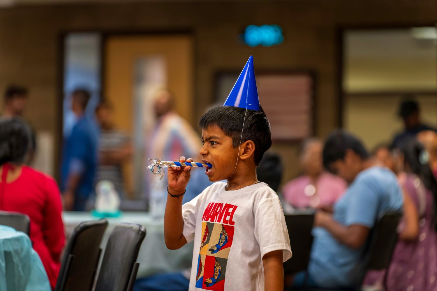 A young boy wearing a Marvel shirt and blue party hat blows a party horn at a celebration.