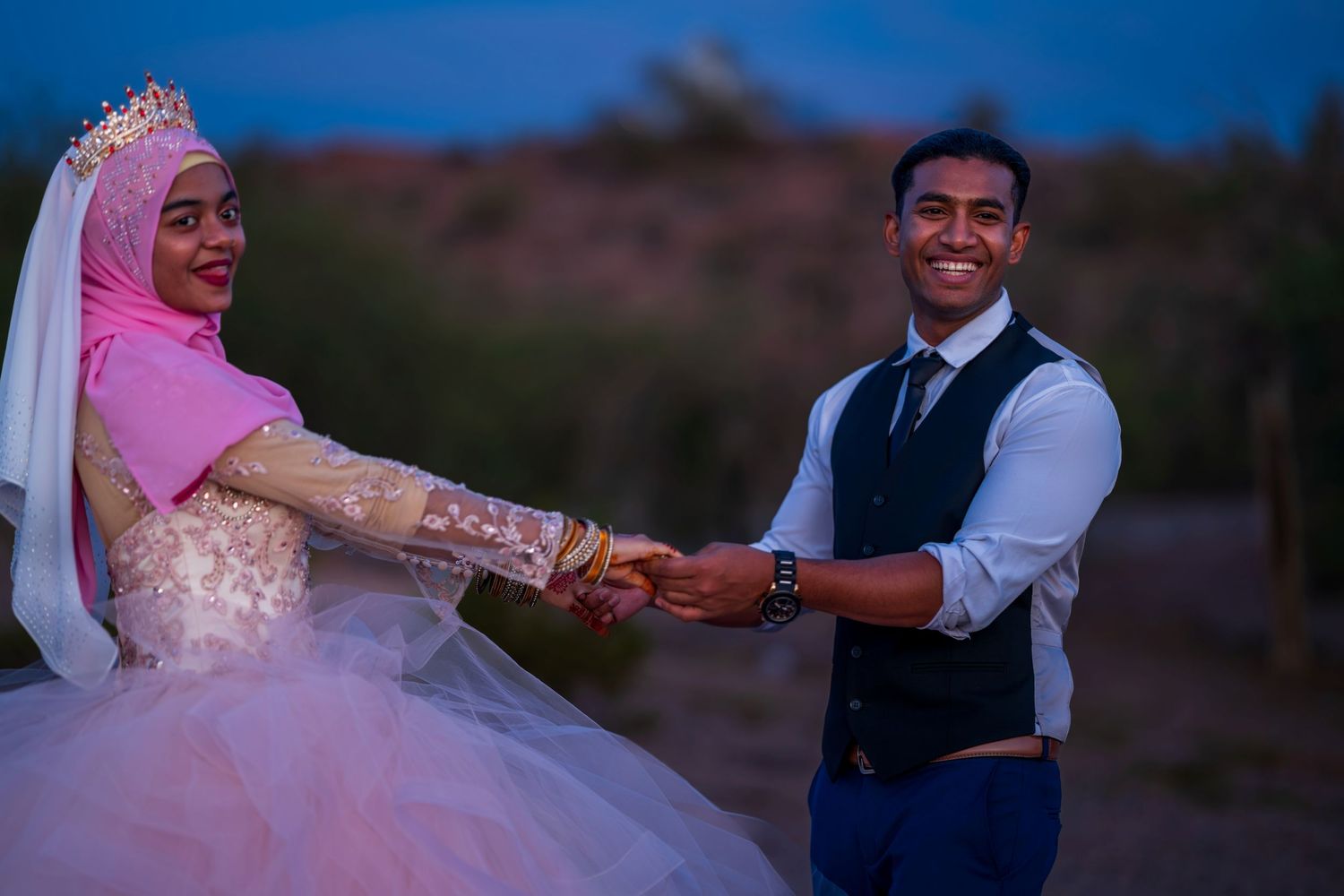 Muslim bride in pink hijab and groom in navy vest hold hands during outdoor desert wedding photoshoot.