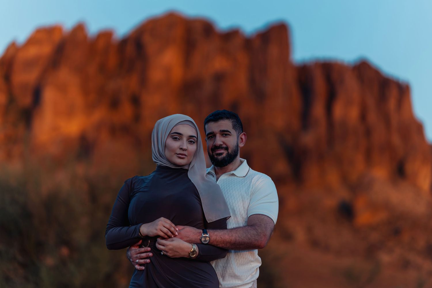 Couple poses together at sunset with dramatic red rock mountain formation in background.