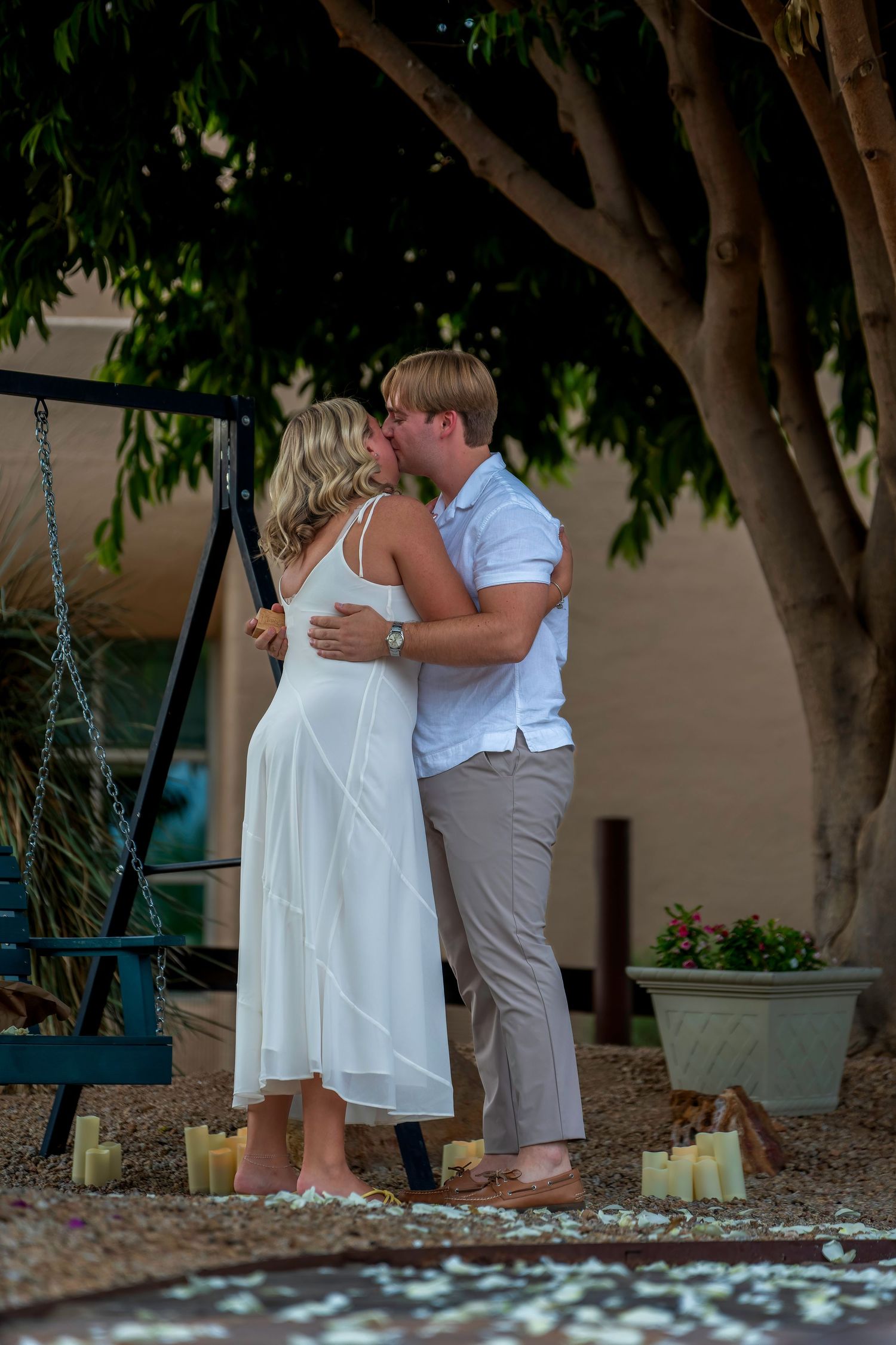 Couple sharing a romantic kiss under a tree surrounded by candles and rose petals during an outdoor proposal.
