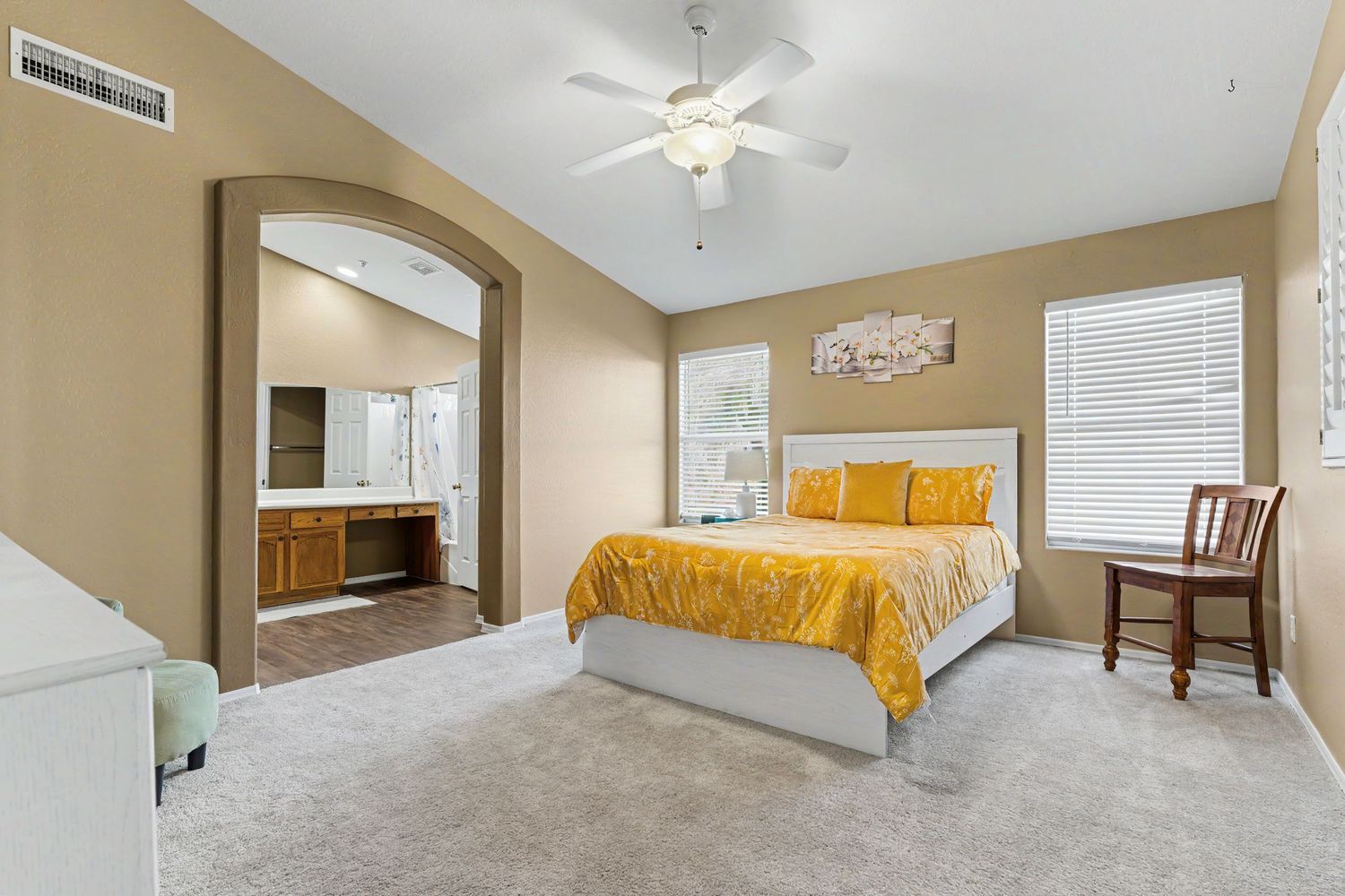 Spacious master bedroom with yellow bedding, ceiling fan, plush carpet, and an arched doorway leading to an en-suite bathroom.