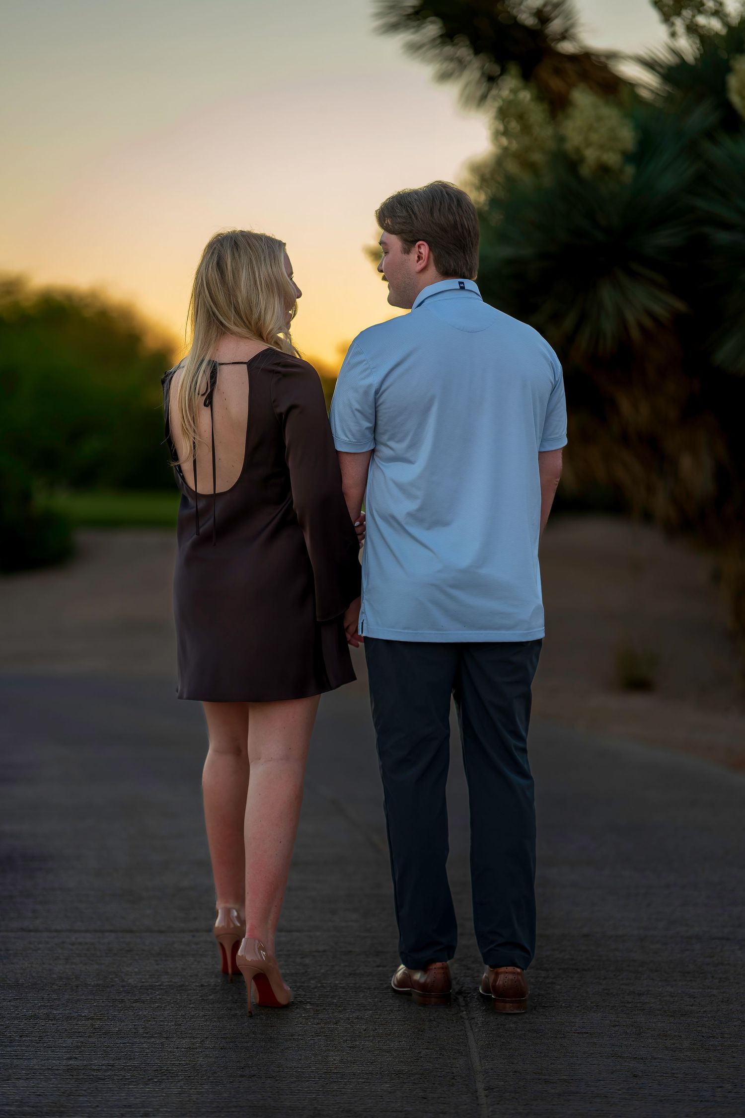 Couple holding hands walking at sunset on a tree-lined path, woman in brown dress and red heels.