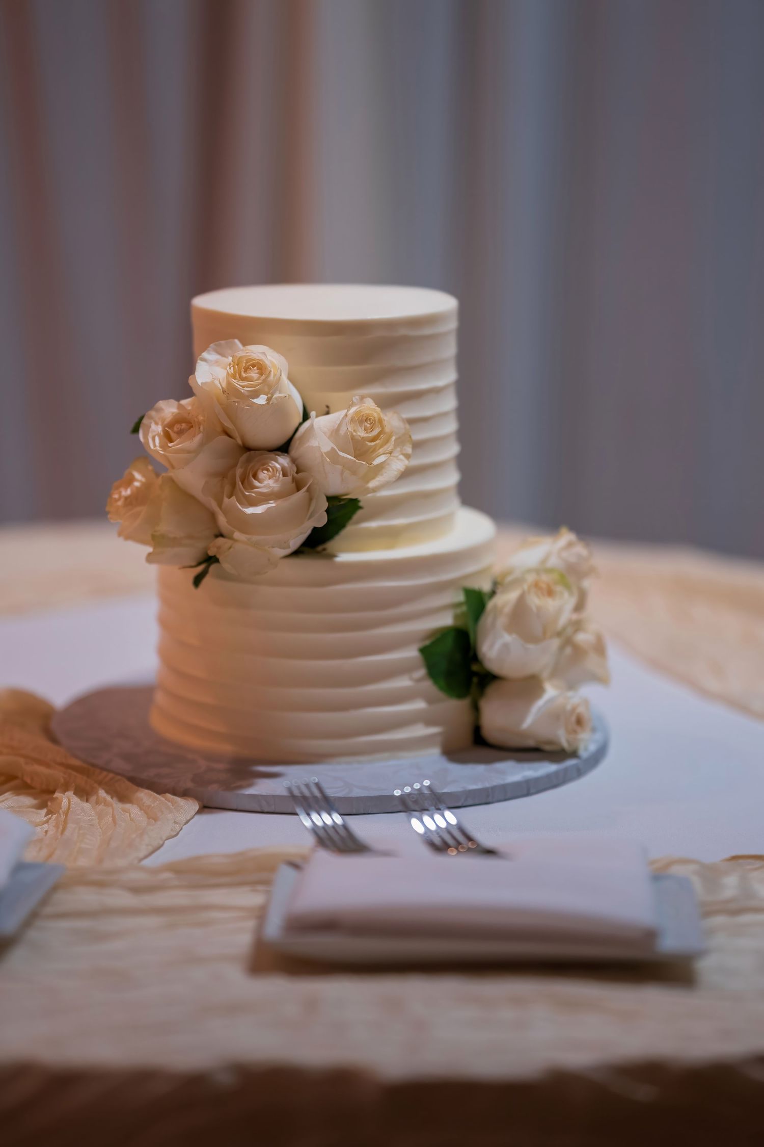 White wedding cake decorated with delicate cream roses sits on an elegant table setting.