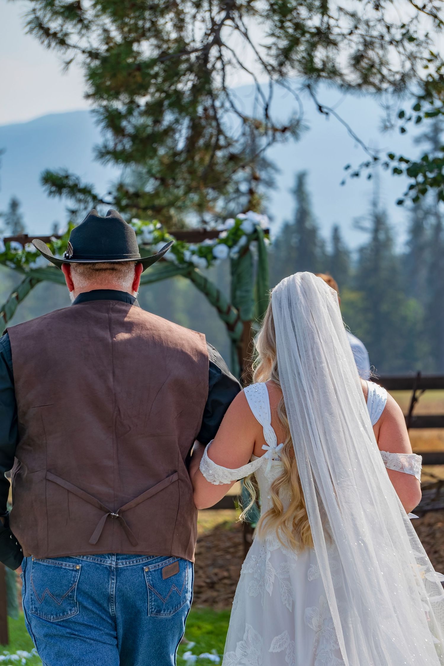 A wedding couple walks together on a grassy outdoor path with pine trees in the background.