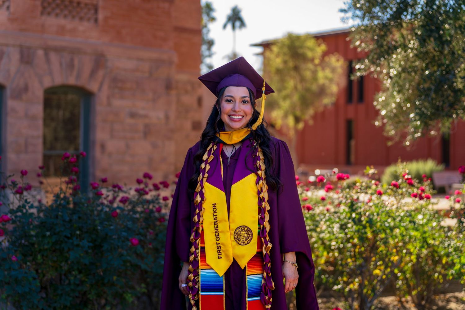 Graduate in academic regalia posing in front of brick building with flowering garden.