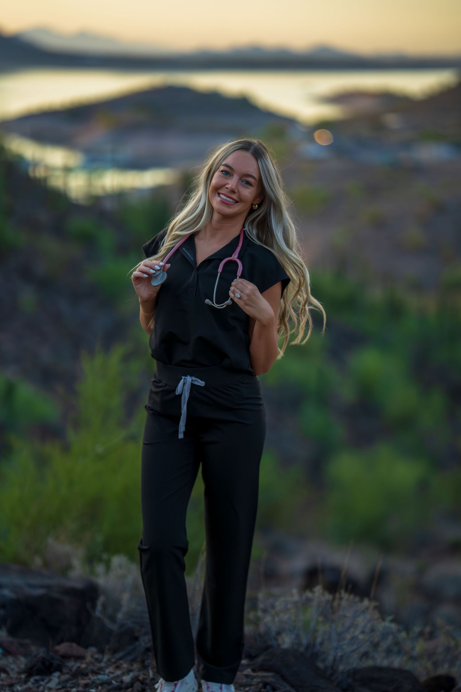 Healthcare worker with stethoscope poses cheerfully against scenic mountain vista at sunset.