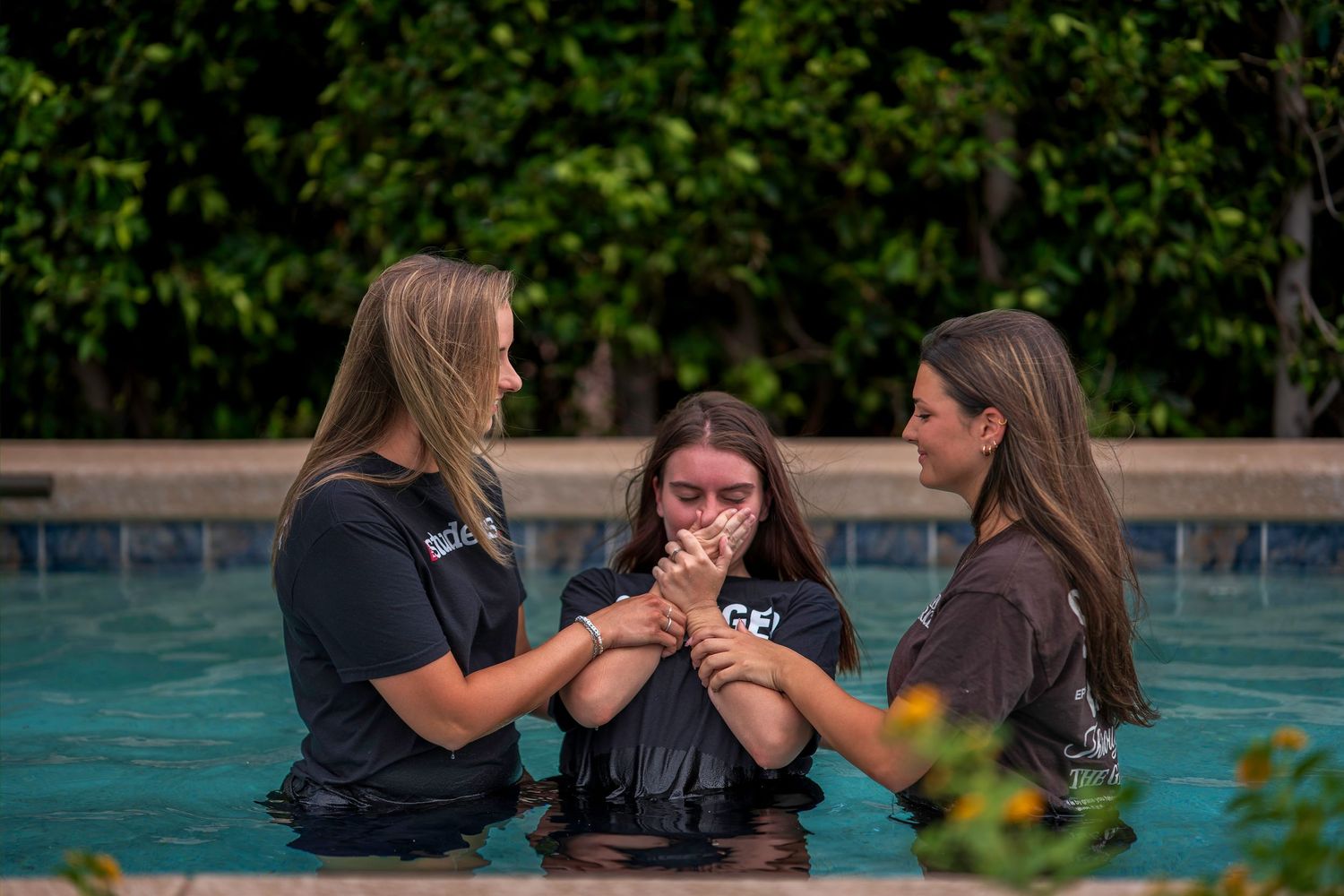 Three women standing in a pool during a baptism ceremony, holding hands in prayer together.