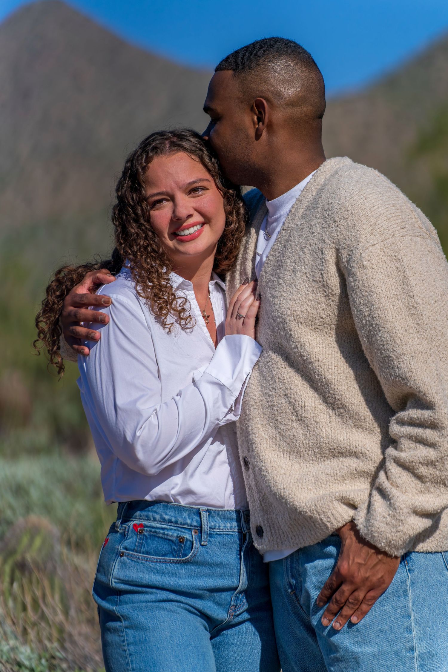 Couple in casual attire embracing against desert mountain backdrop.