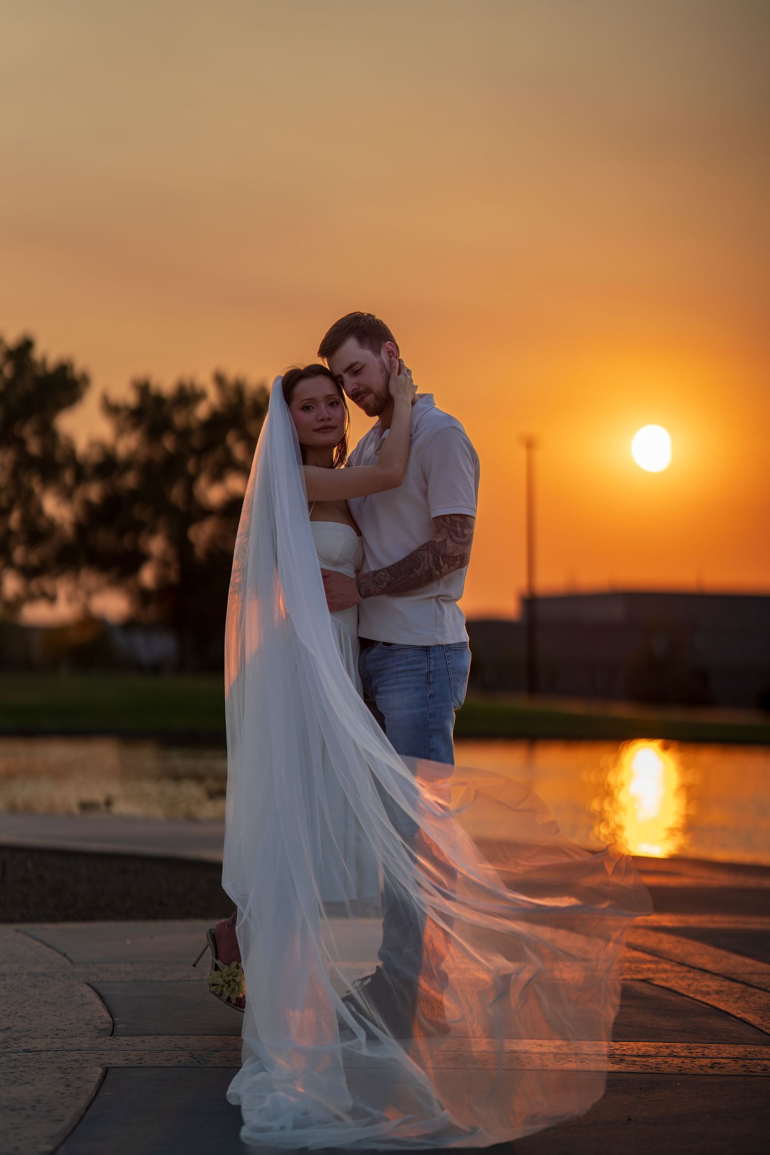 A couple embraces during a romantic sunset photoshoot by the water with a flowing white veil catching the breeze.