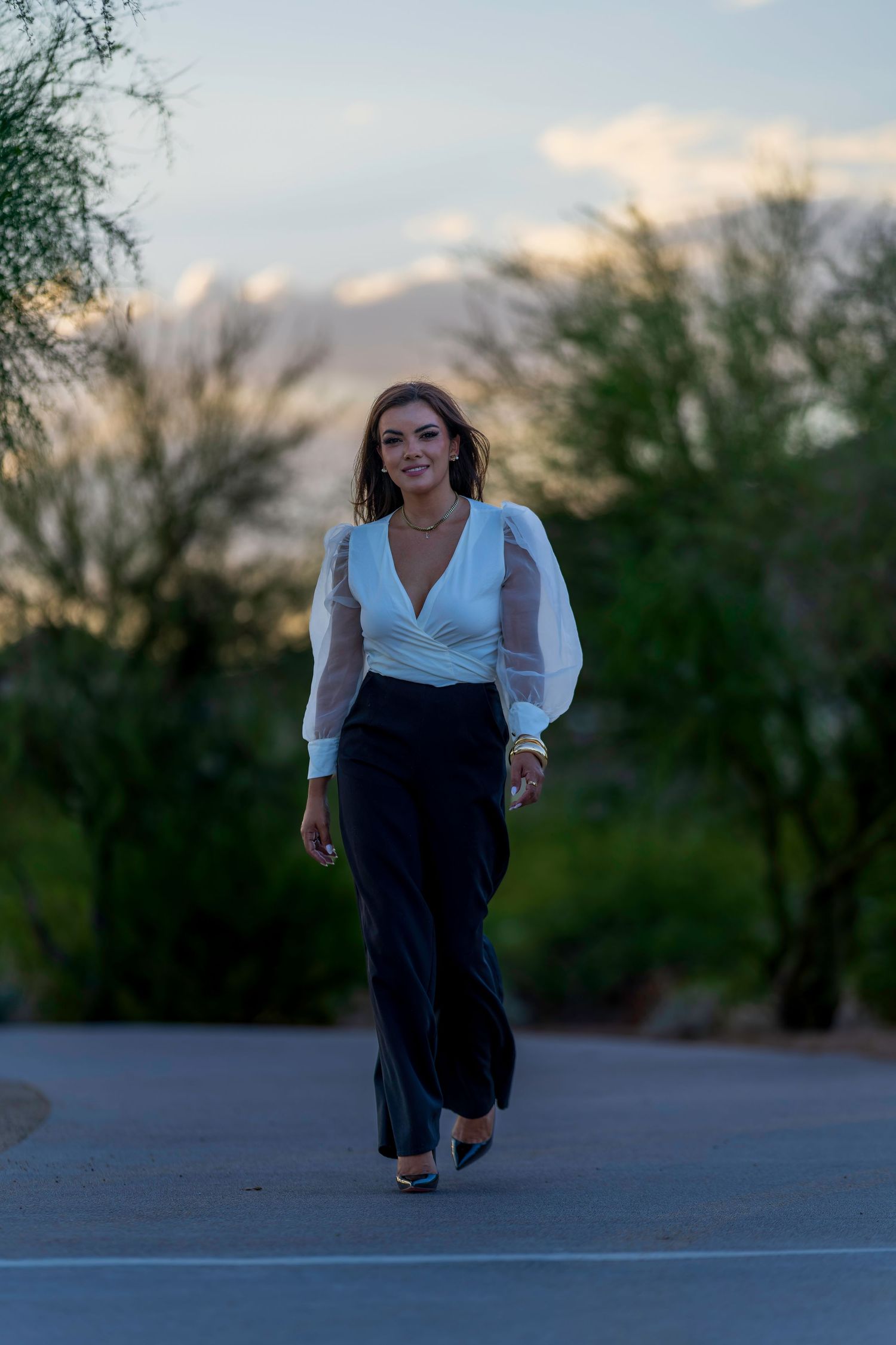 Smiling woman walking in white sheer-sleeve top and black wide-leg trousers on outdoor path at dusk.