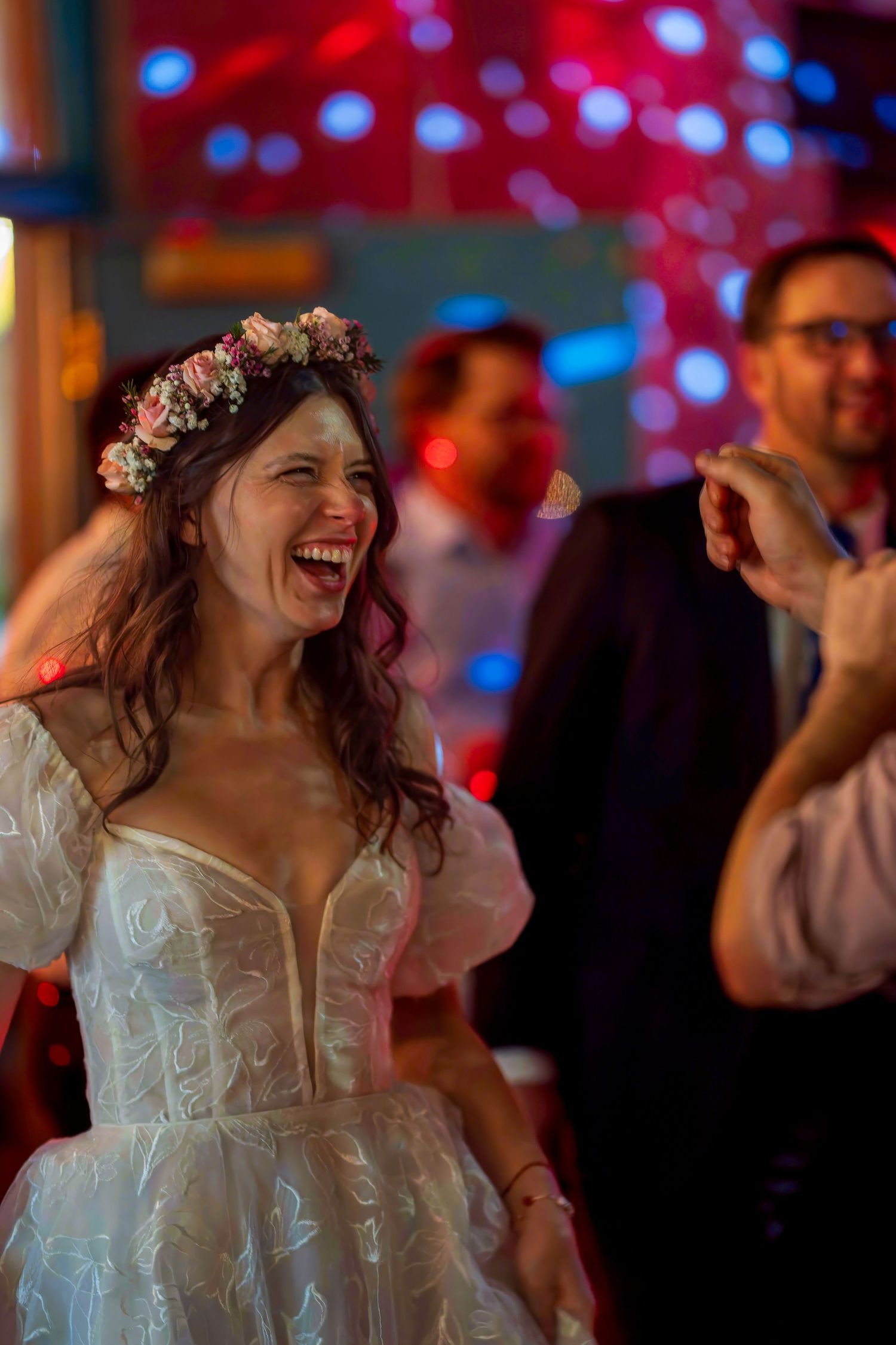 Joyful bride in white lace dress and floral crown laughing while dancing at wedding reception.