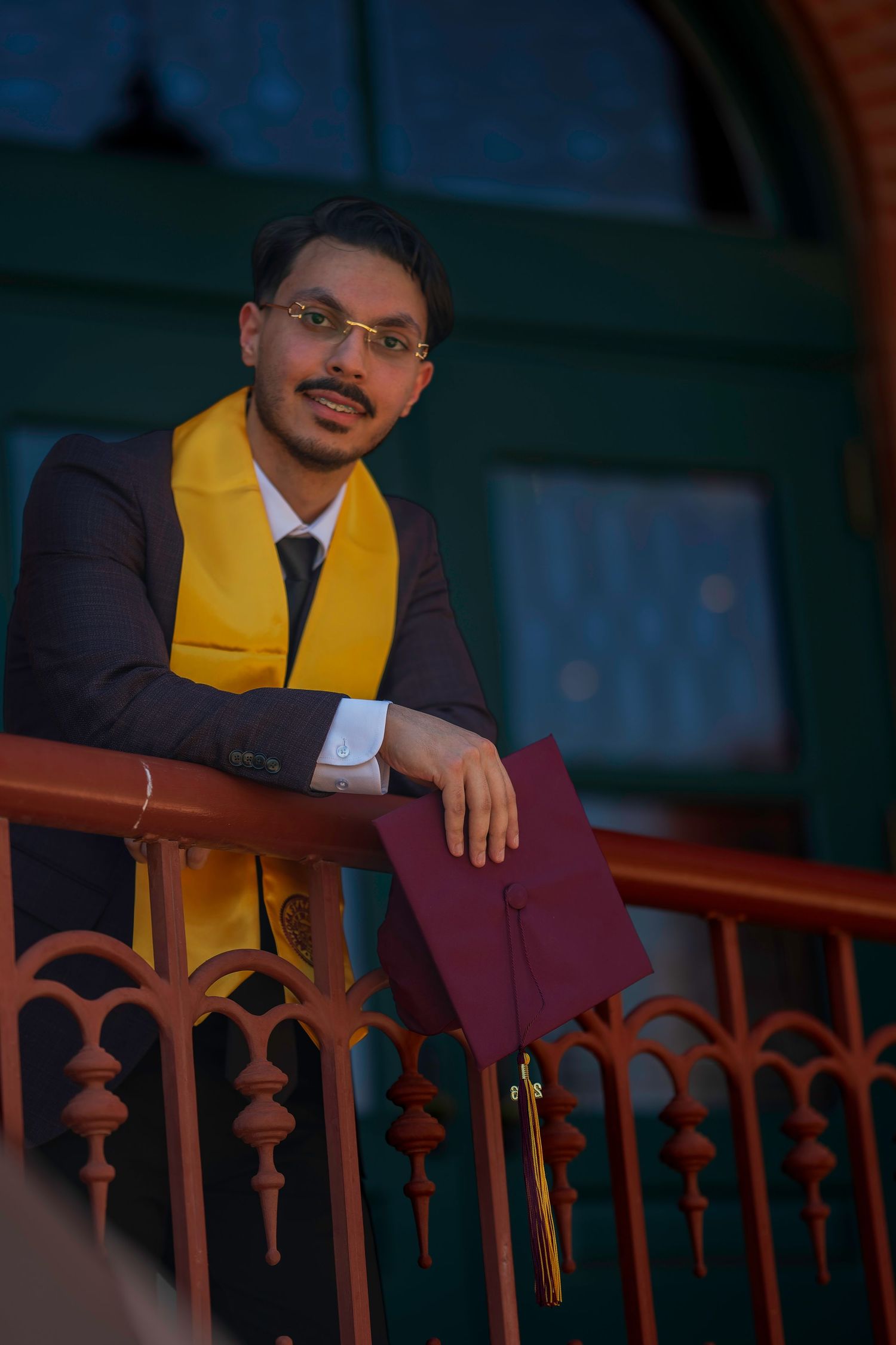 Young graduate in dark suit with yellow stole holds maroon cap while leaning on ornate railing.
