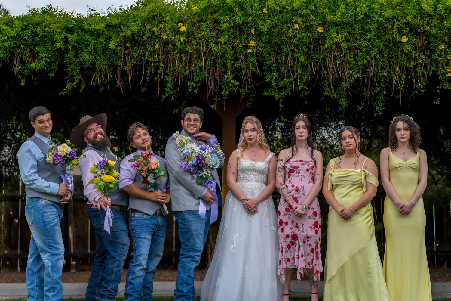 Wedding party poses outdoors with groomsmen in jeans holding bouquets beside bride and bridesmaids in floral and yellow gowns.