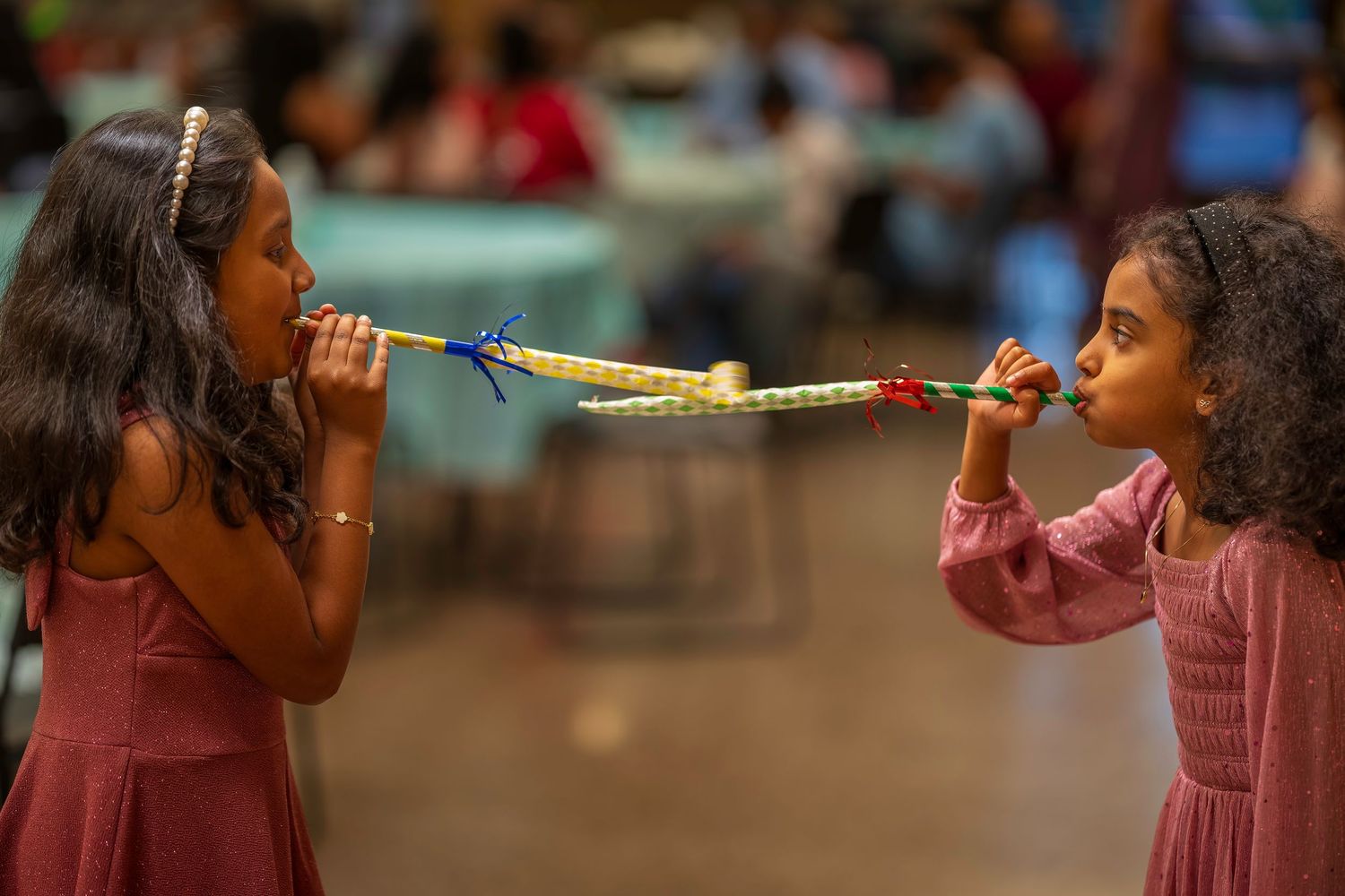 Two young girls in pink dresses blowing into opposite ends of a colorful party horn at a celebration.