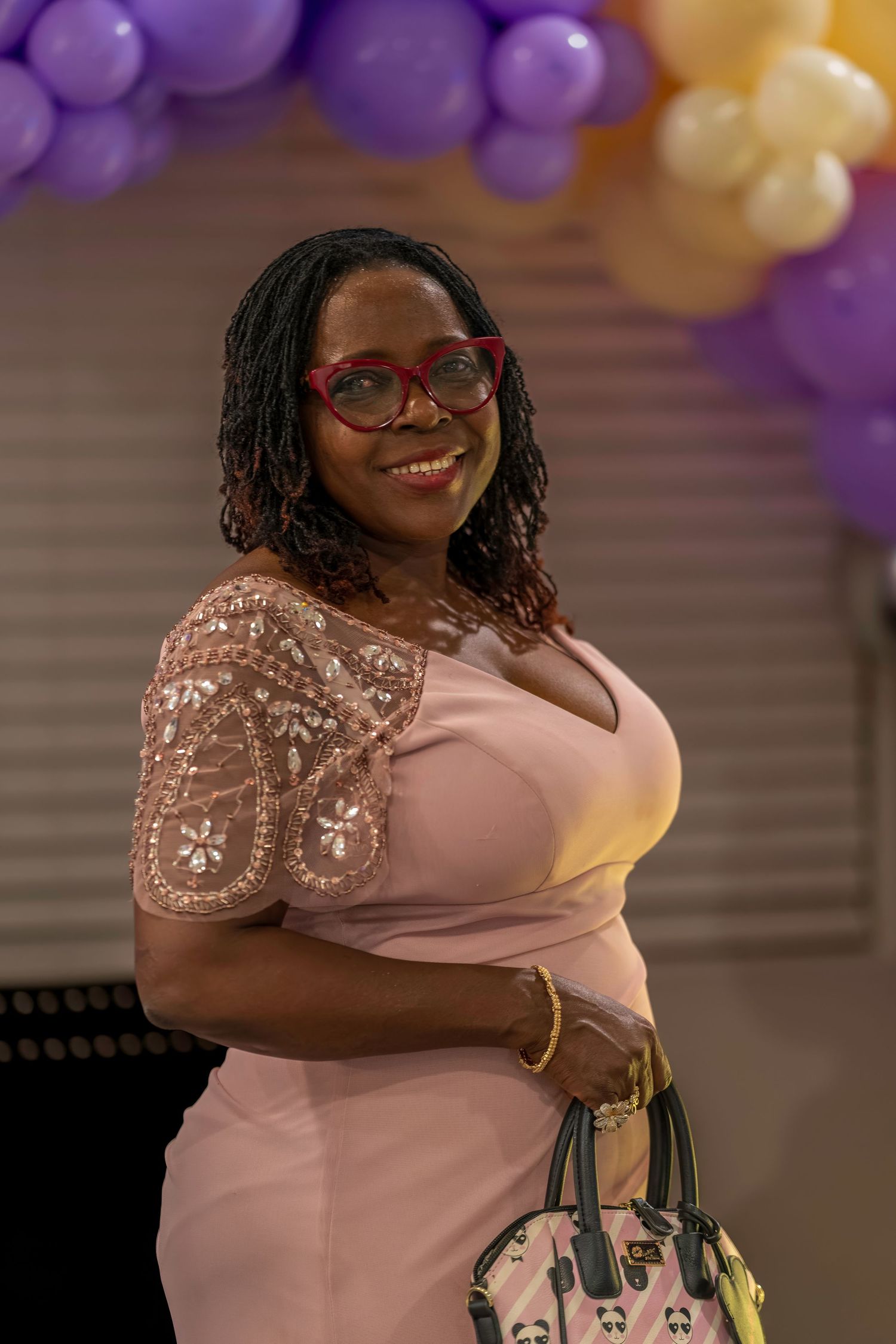 Smiling Black woman in pink lace dress with red glasses holding a patterned purse under colorful balloon arch.