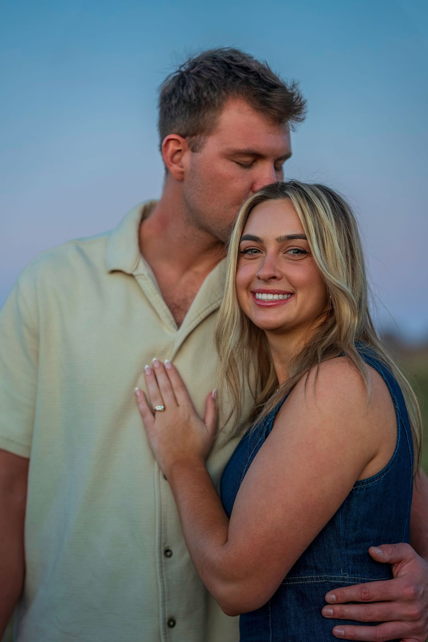 A man kisses a smiling blonde woman wearing an engagement ring outdoors at dusk.