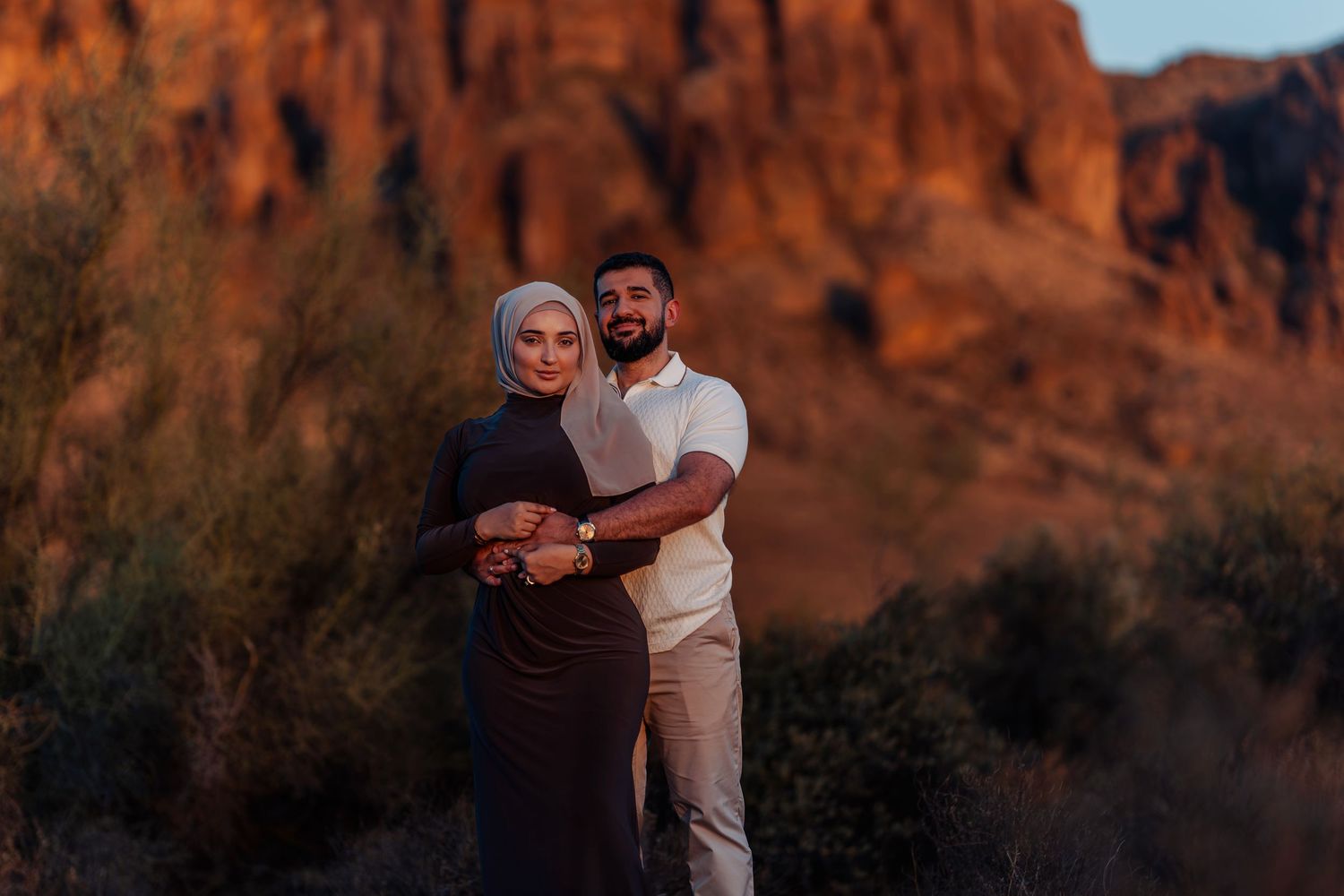 A couple poses together at sunset in the desert, with stunning red rock cliffs glowing behind them.