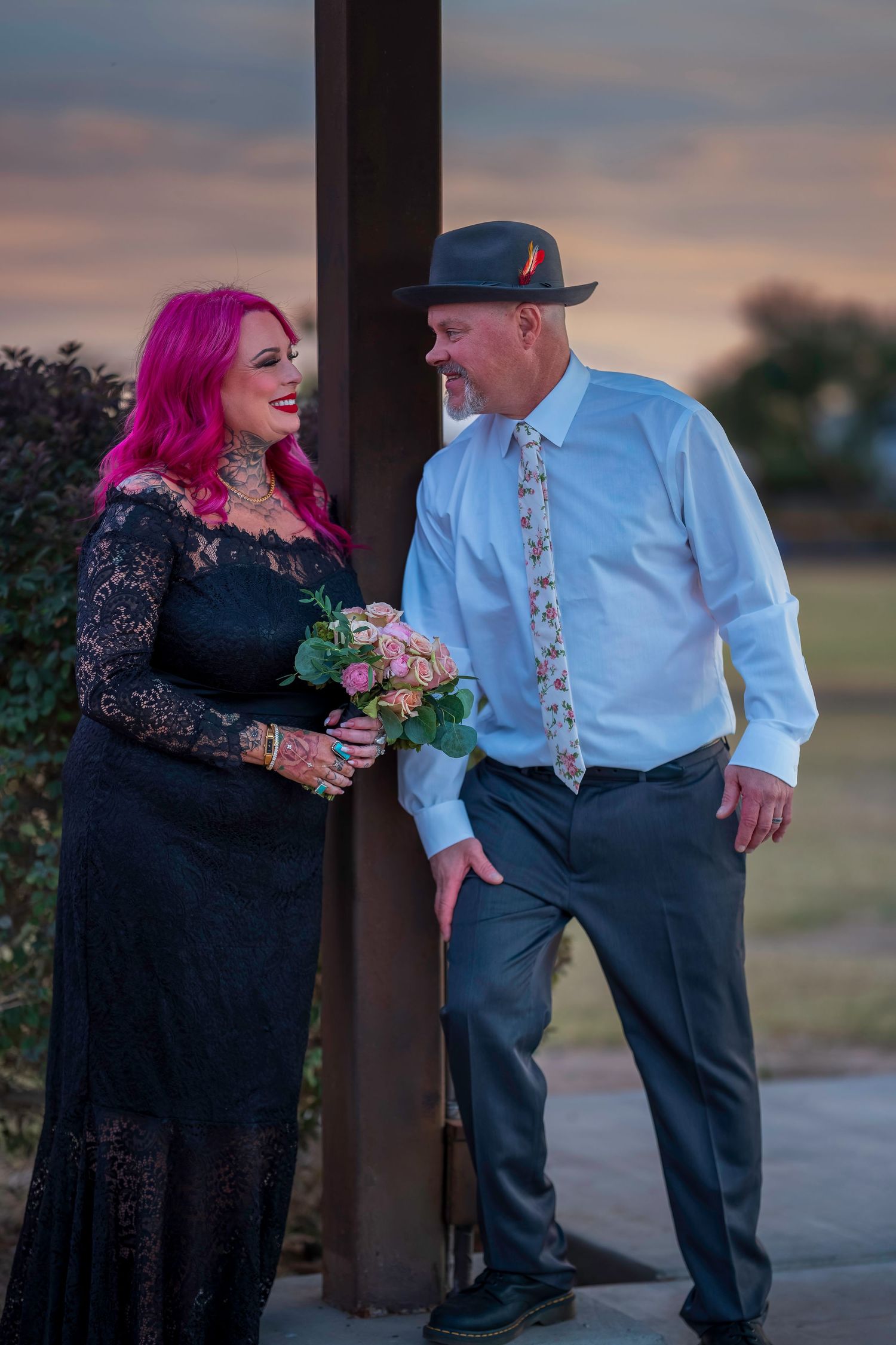Couple in formal attire, woman with pink hair in black lace dress, man in hat and floral tie, holding bouquet at sunset.