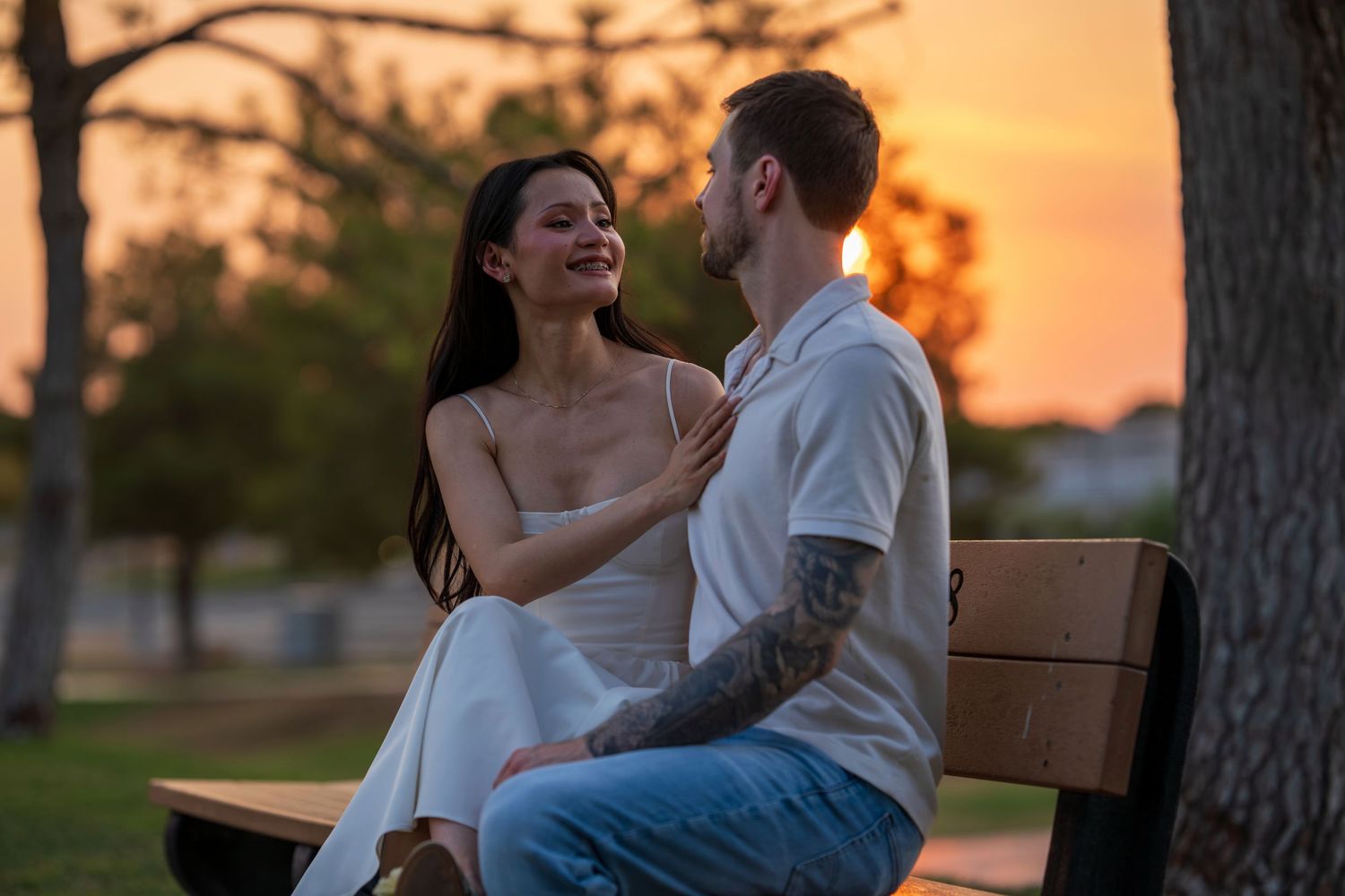 A smiling couple sits on a park bench at sunset sharing a tender moment.