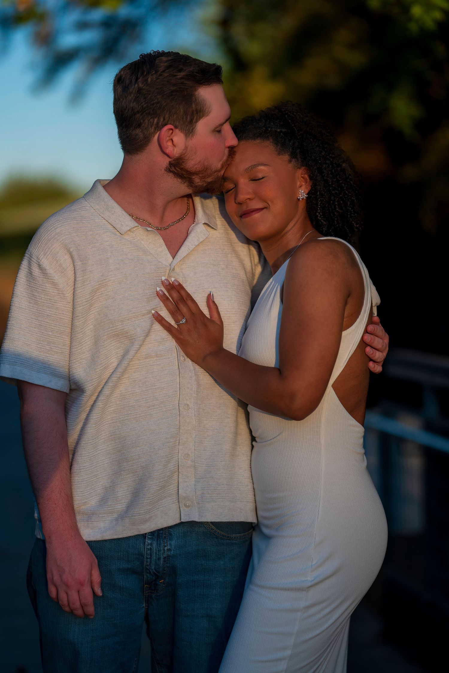 Romantic interracial couple embracing outdoors at golden hour, woman showing engagement ring in white dress.